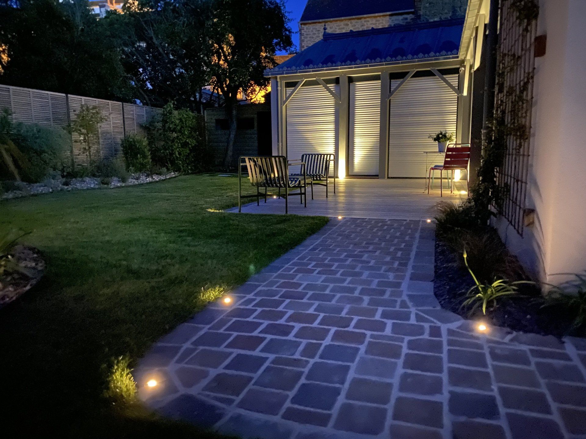 Allée de terrasse en pierre éclairée par des spots au sol menant à un gazebo de jardin chaleureusement éclairé au crépuscule.