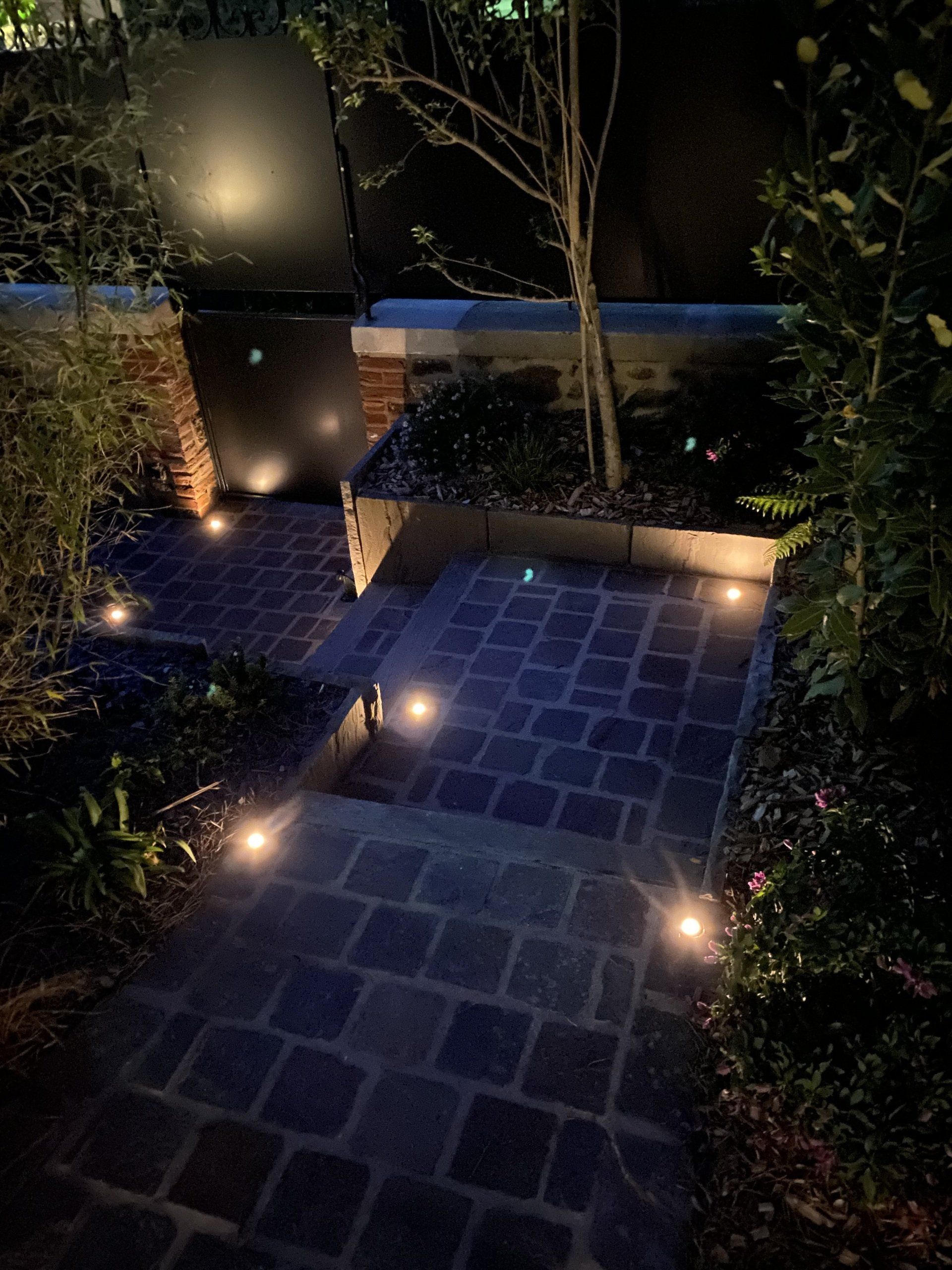 Allée nocturne du jardin avec de petits éclairages au sol, allée carrelée et arbres bordant un point d'eau doucement éclairé.