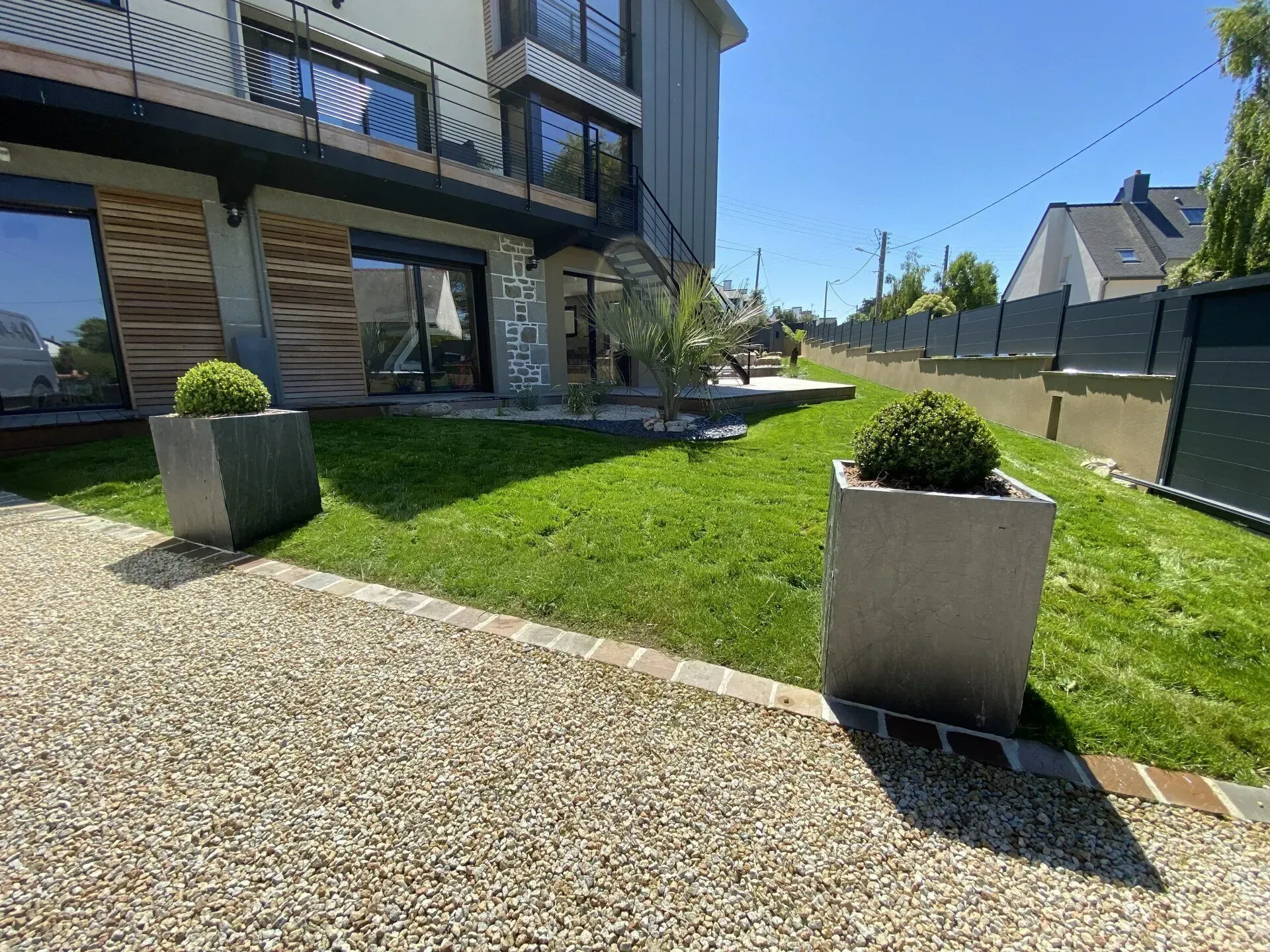 Terrasse et pelouse d'une maison moderne avec deux jardinières carrées sous un ciel d'un bleu éclatant.