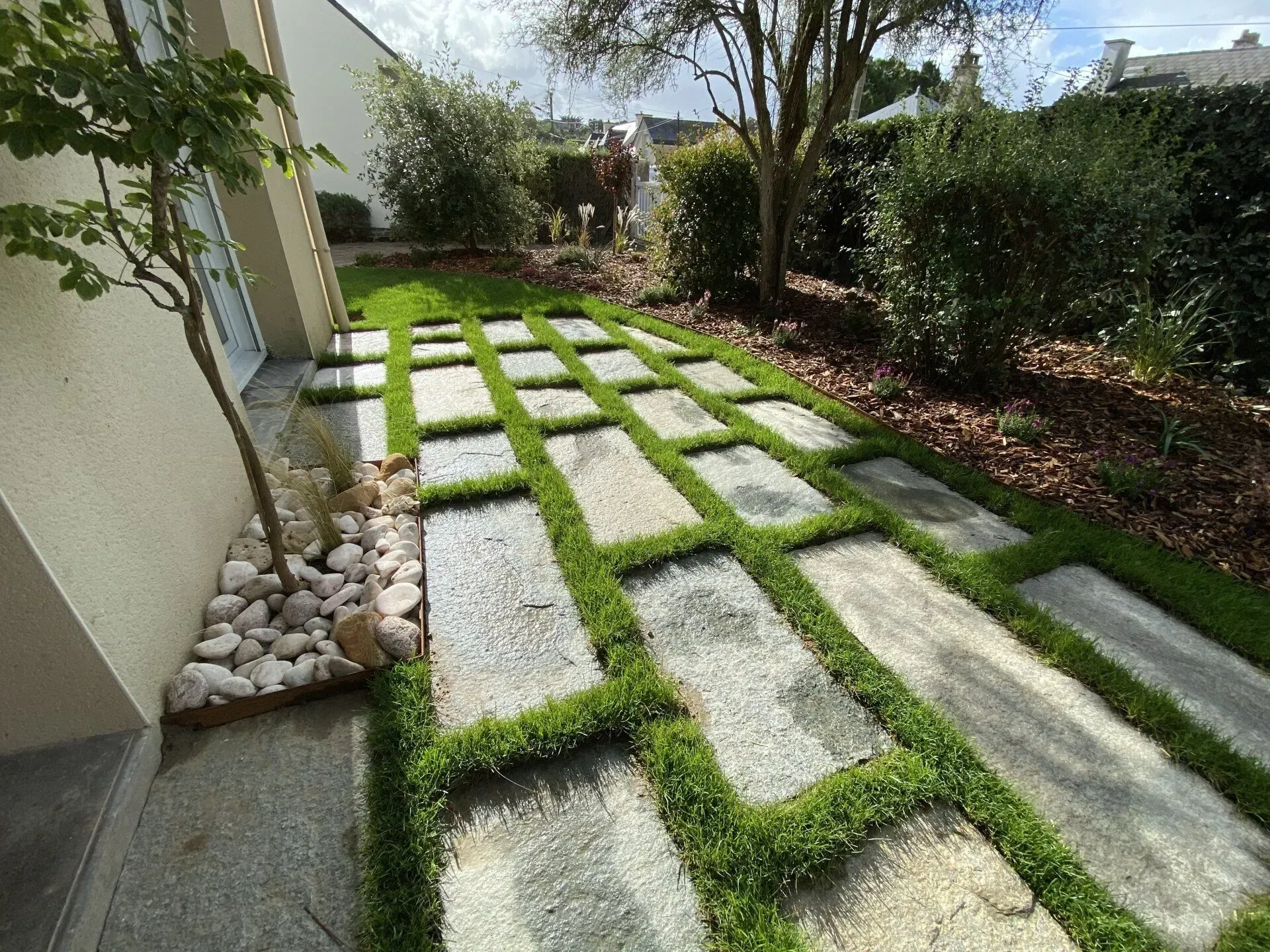 Allée de jardin moderne avec de grandes dalles, gazon vert entre les pavés et arbustes le long d'un mur de maison.