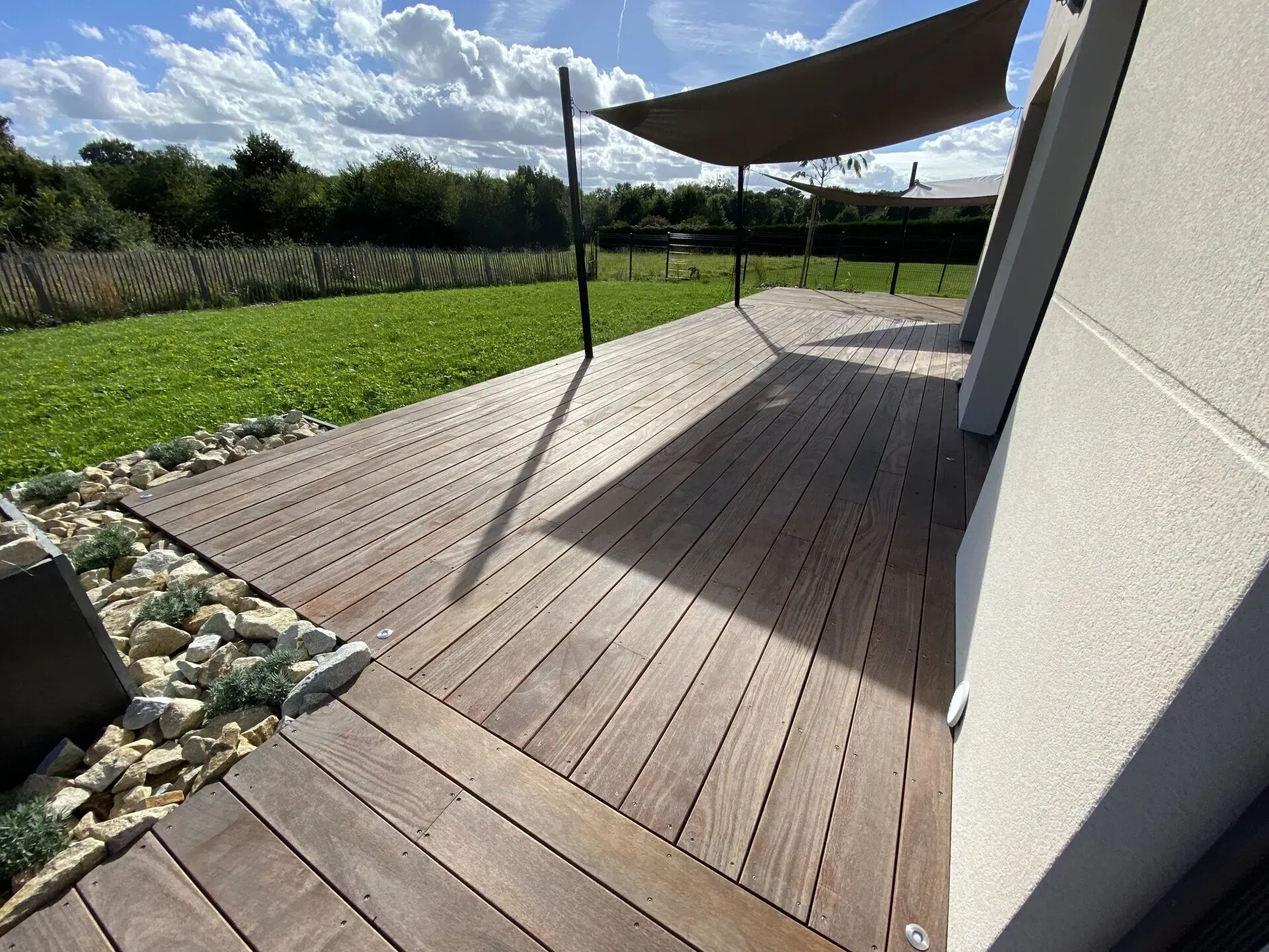 Terrasse moderne en bois jouxtant une maison blanche, avec une voile d'ombrage au-dessus d'une cour gazonnée sous un ciel nuageux.