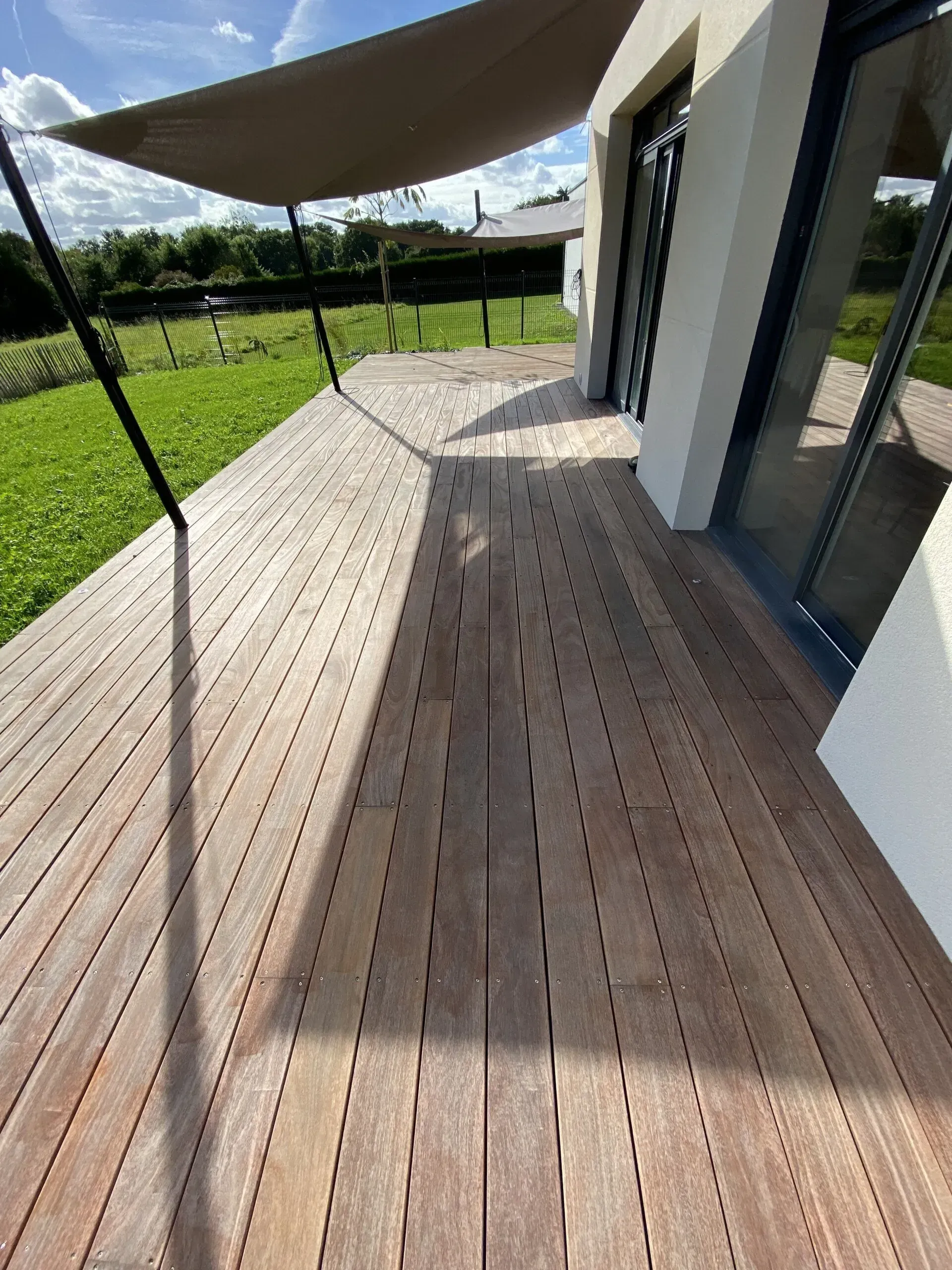 Terrasse en bois jouxtant une maison moderne, avec une voile d'ombrage au-dessus et une pelouse verte au-delà.
