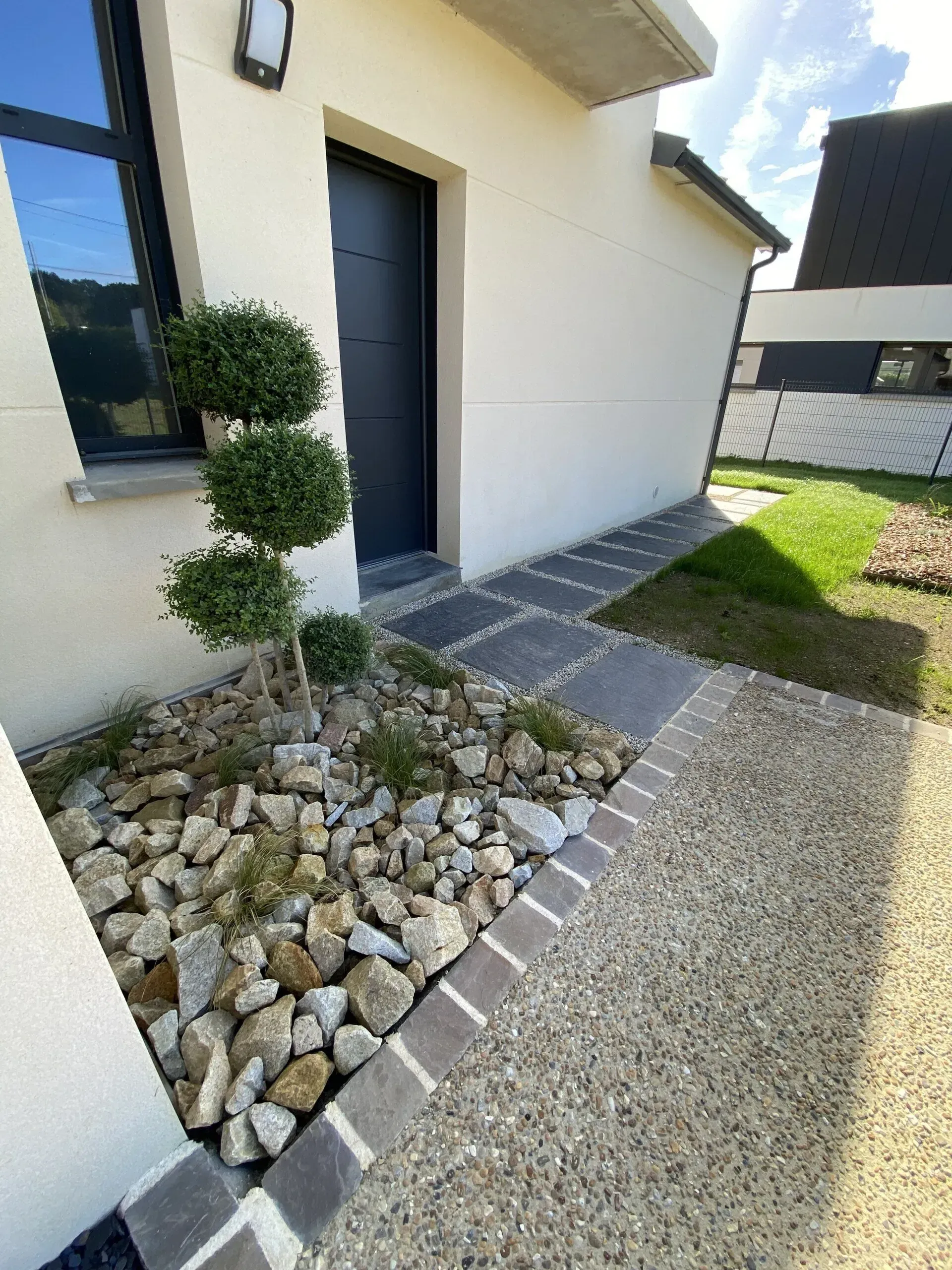 Entrée de maison moderne avec chemin de gravier, bordure en pierre et petit arbuste topiaire près d'une porte ouverte.