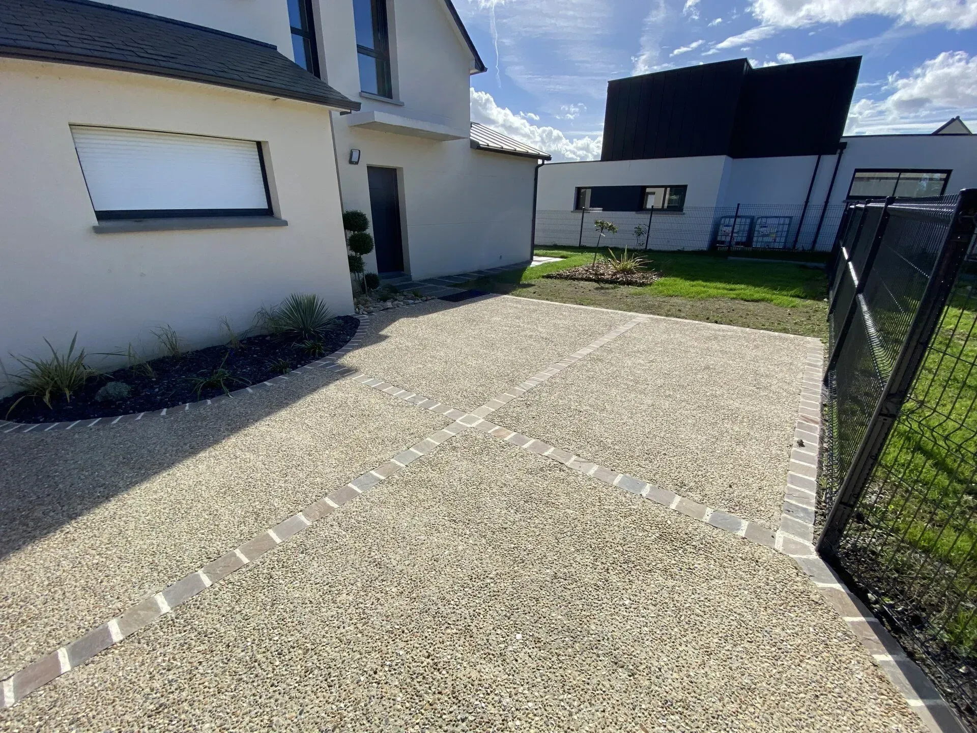 Allée de gravier longeant une maison blanche, avec un portail noir et une petite pelouse en arrière-plan.