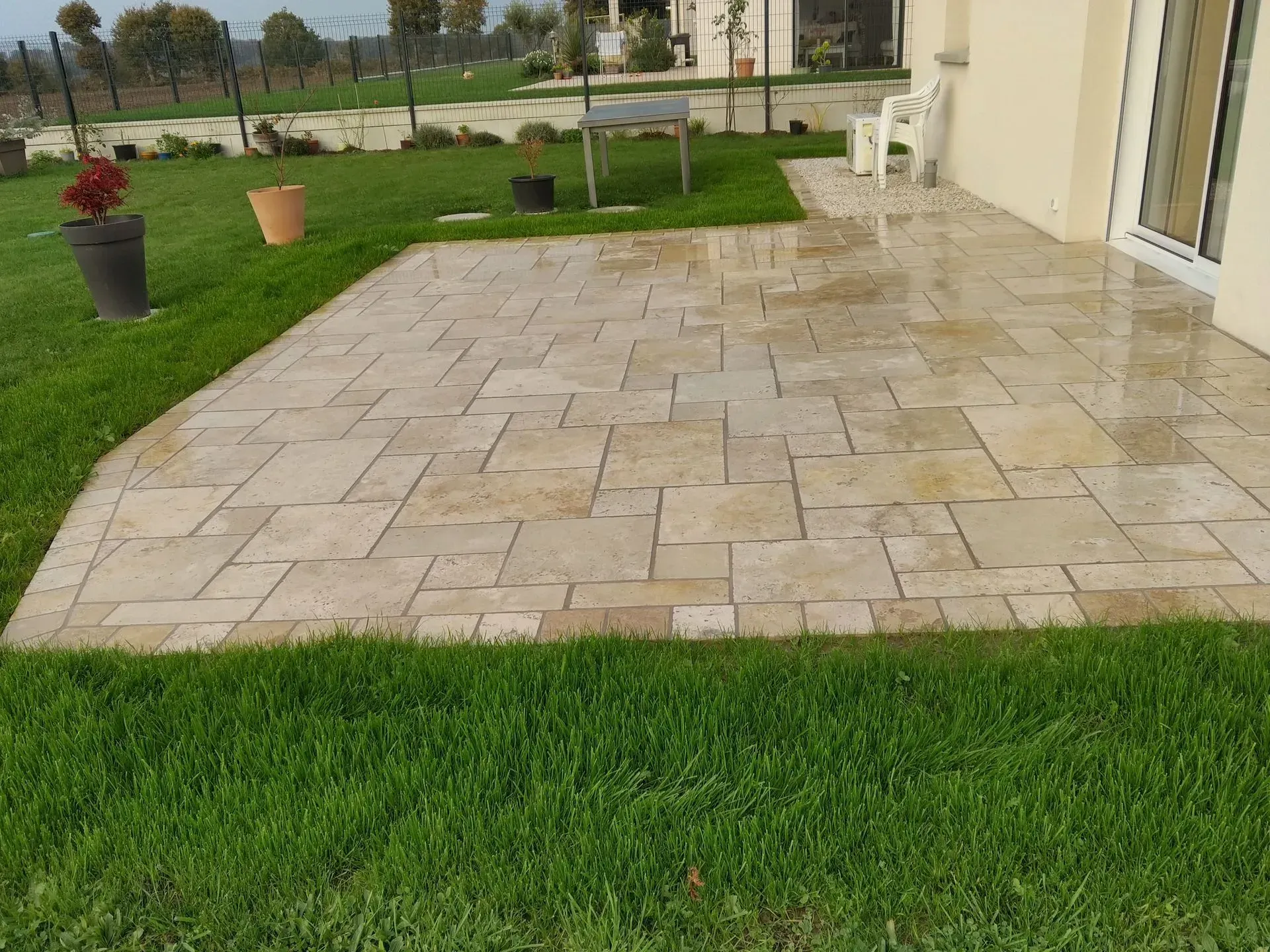 Terrasse pavée de dalles claires, bordée par une pelouse et une maison, avec des plantes en pot en arrière-plan.