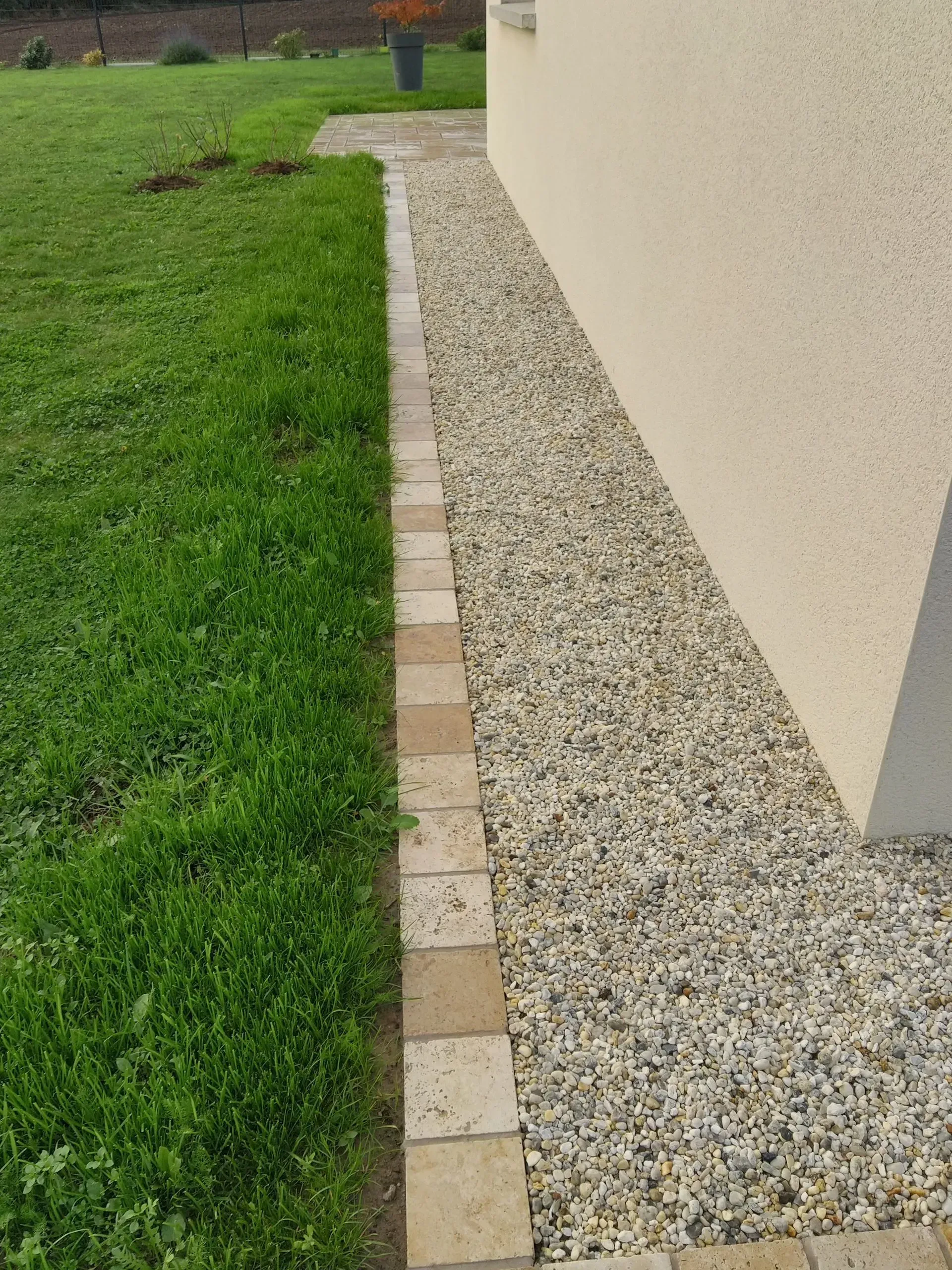 Bordure de gravier le long d'un mur beige, avec une pelouse verte et une étroite bande de pavés en bordure.