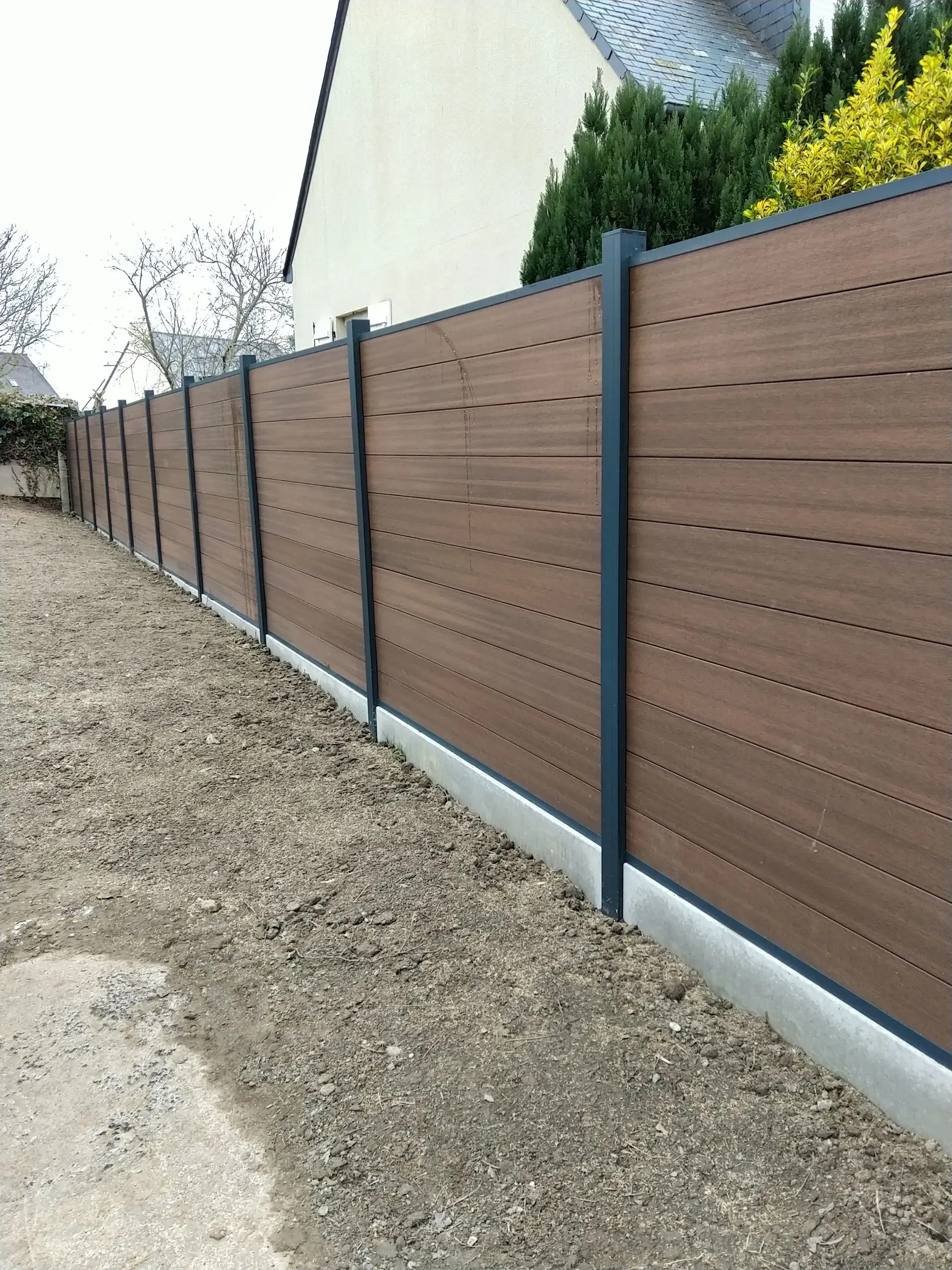 Clôture en bois brun pour préserver l'intimité le long d'un chemin de gravier, à côté d'une maison et d'arbustes.
