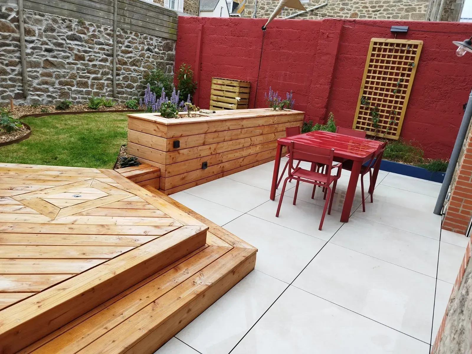 Patio avec table et chaises rouges, terrasse en bois, jardinière en briques et pelouse verte adossée à un mur rouge.