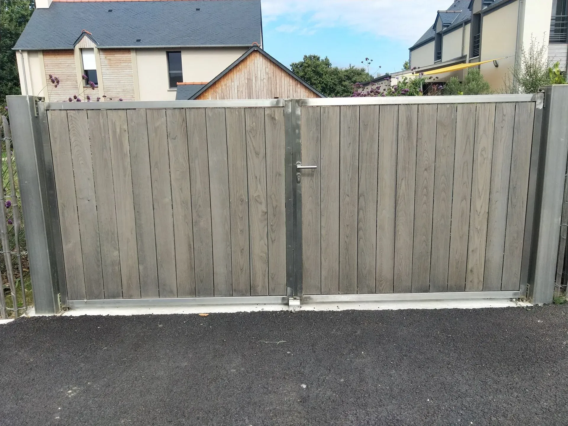 Portail double en bois gris devant une maison, avec une route asphaltée au premier plan.
