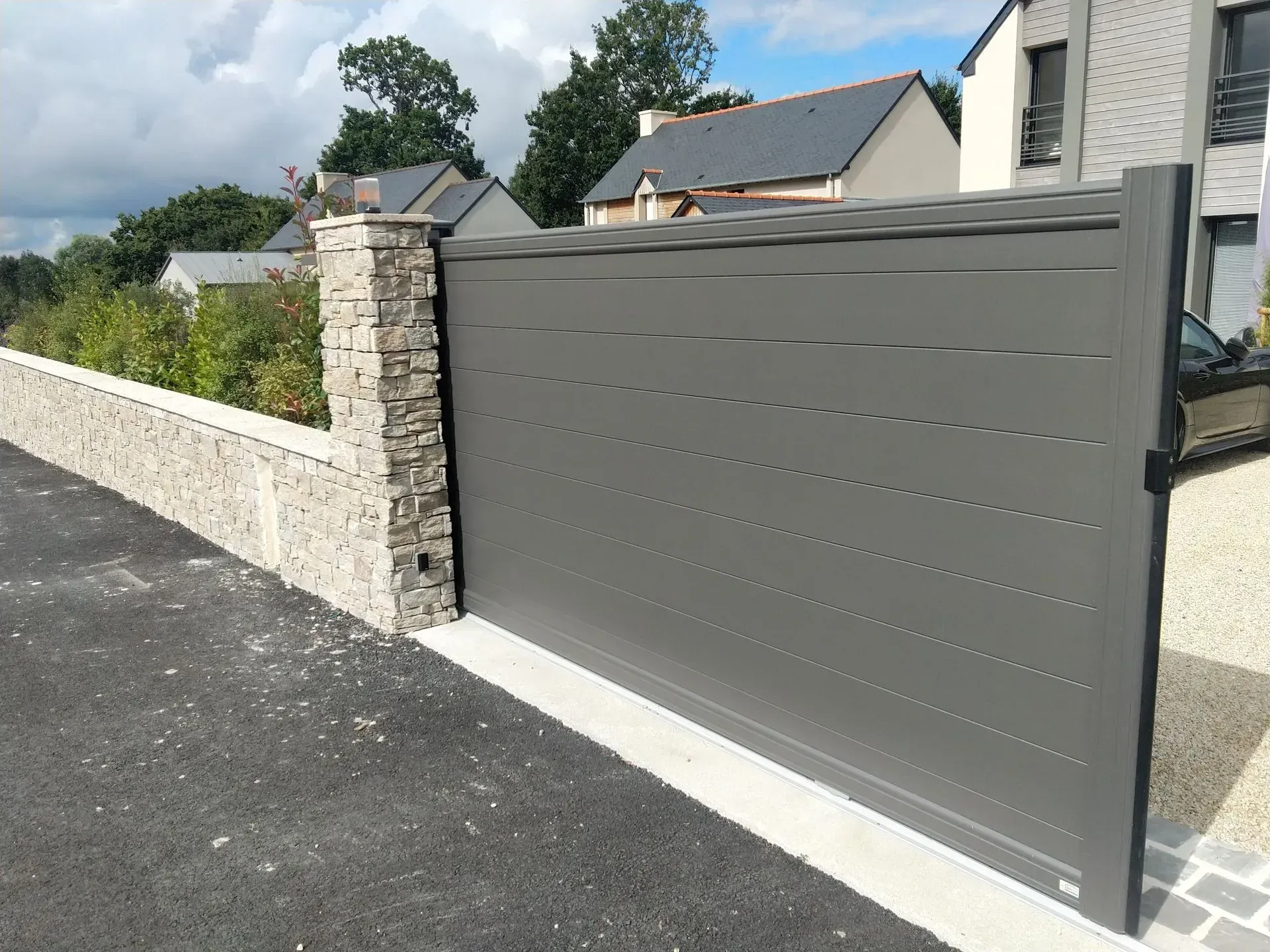 Portail coulissant gris moderne pour allée, jouxtant un mur en pierre et une maison de banlieue.