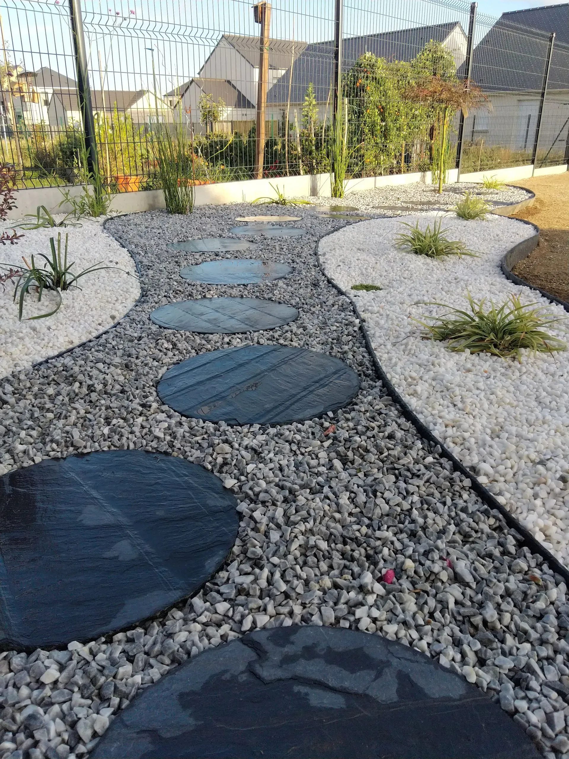 Allée de jardin en gravier avec de grandes dalles noires entre des bordures en pierre claire dans une cour paysagée.