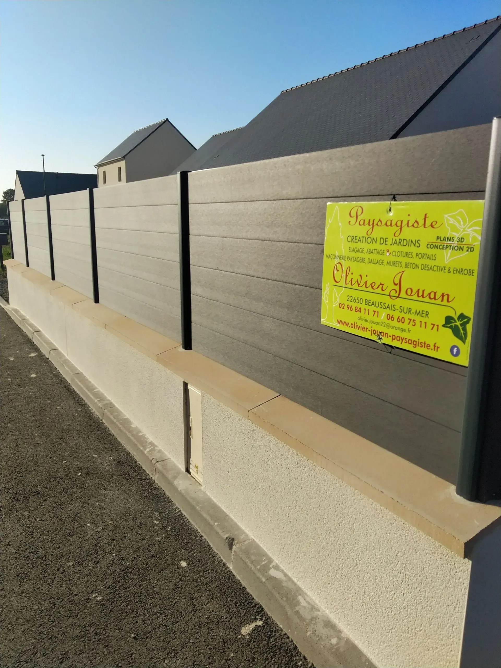 Panneau jaune sur une clôture sombre, au bord d'un chemin de gravier, sous un ciel dégagé.