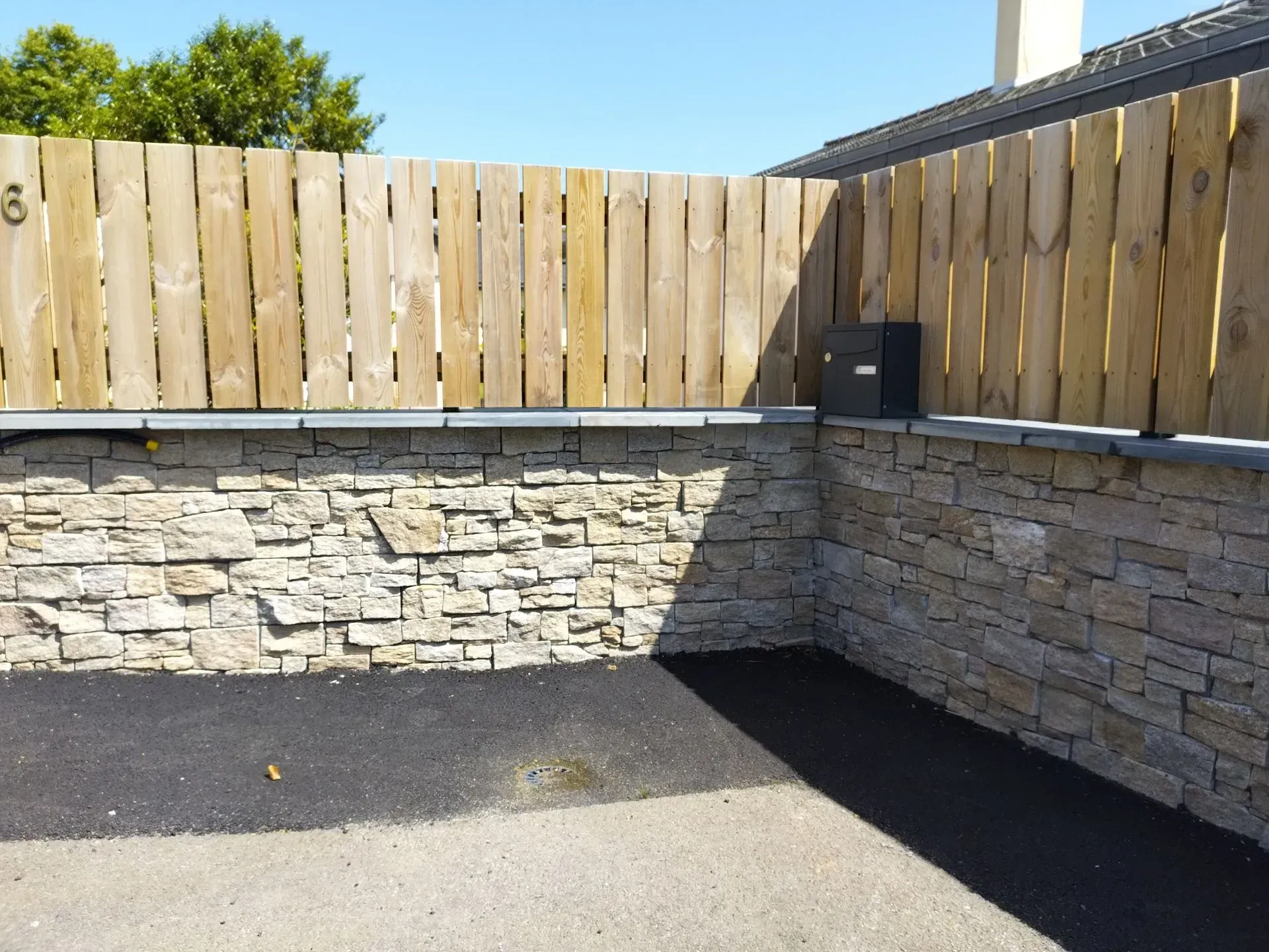Coin de terrasse en pierre avec murets en pierres empilées, clôture en bois et pavage sombre sous un ciel bleu.
