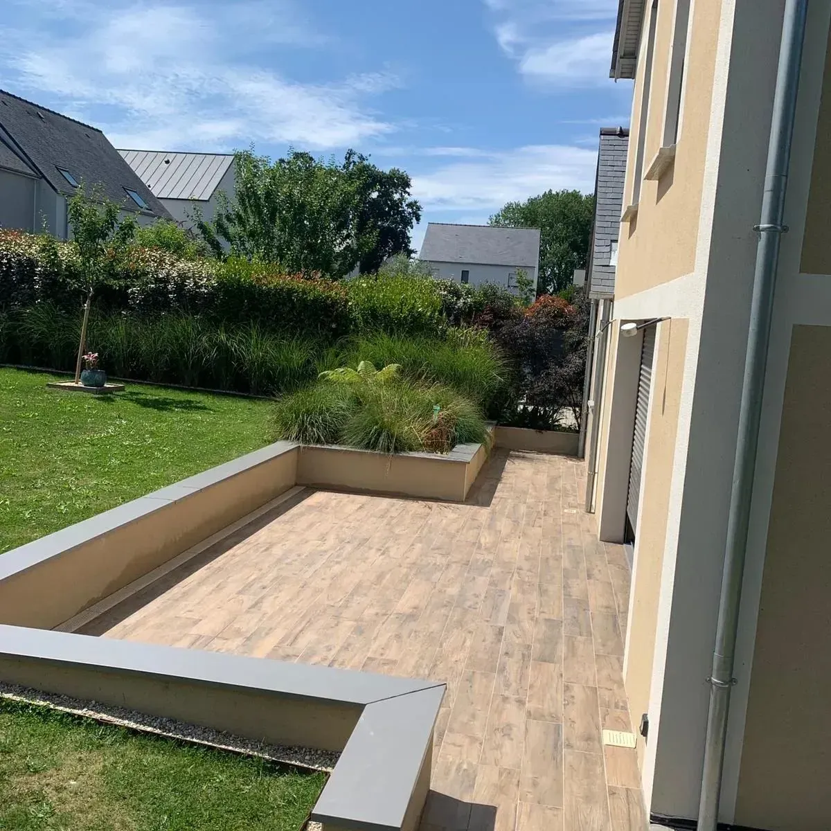 Terrasse ensoleillée à l'arrière avec dalles beiges, parterres de jardin surélevés et pelouse verte à côté d'une maison.