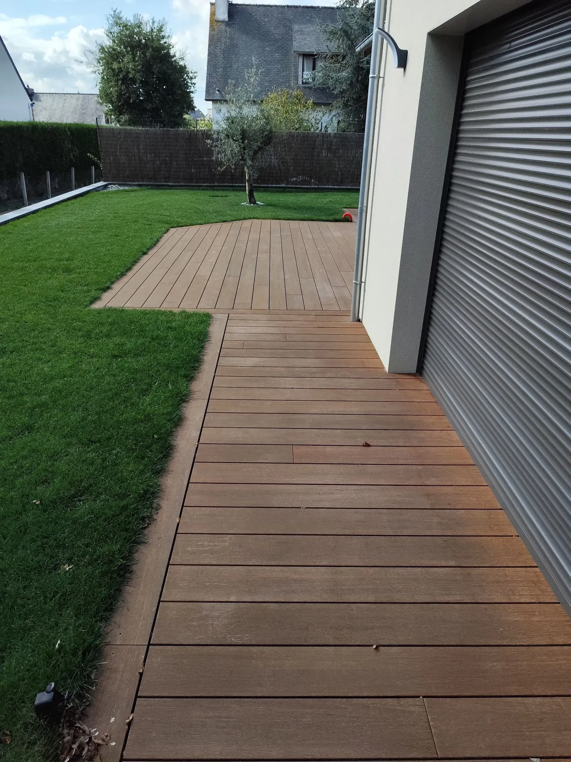 Allée en bois à l'arrière de la maison menant à une petite terrasse, avec pelouse verte et haies.