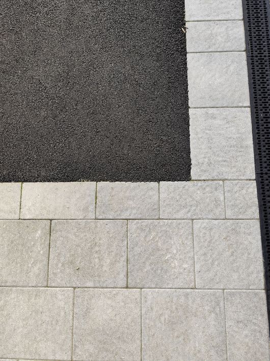 Allée pavée de carreaux gris clair et d'une section en gravier ou en asphalte texturé foncé en haut à gauche.