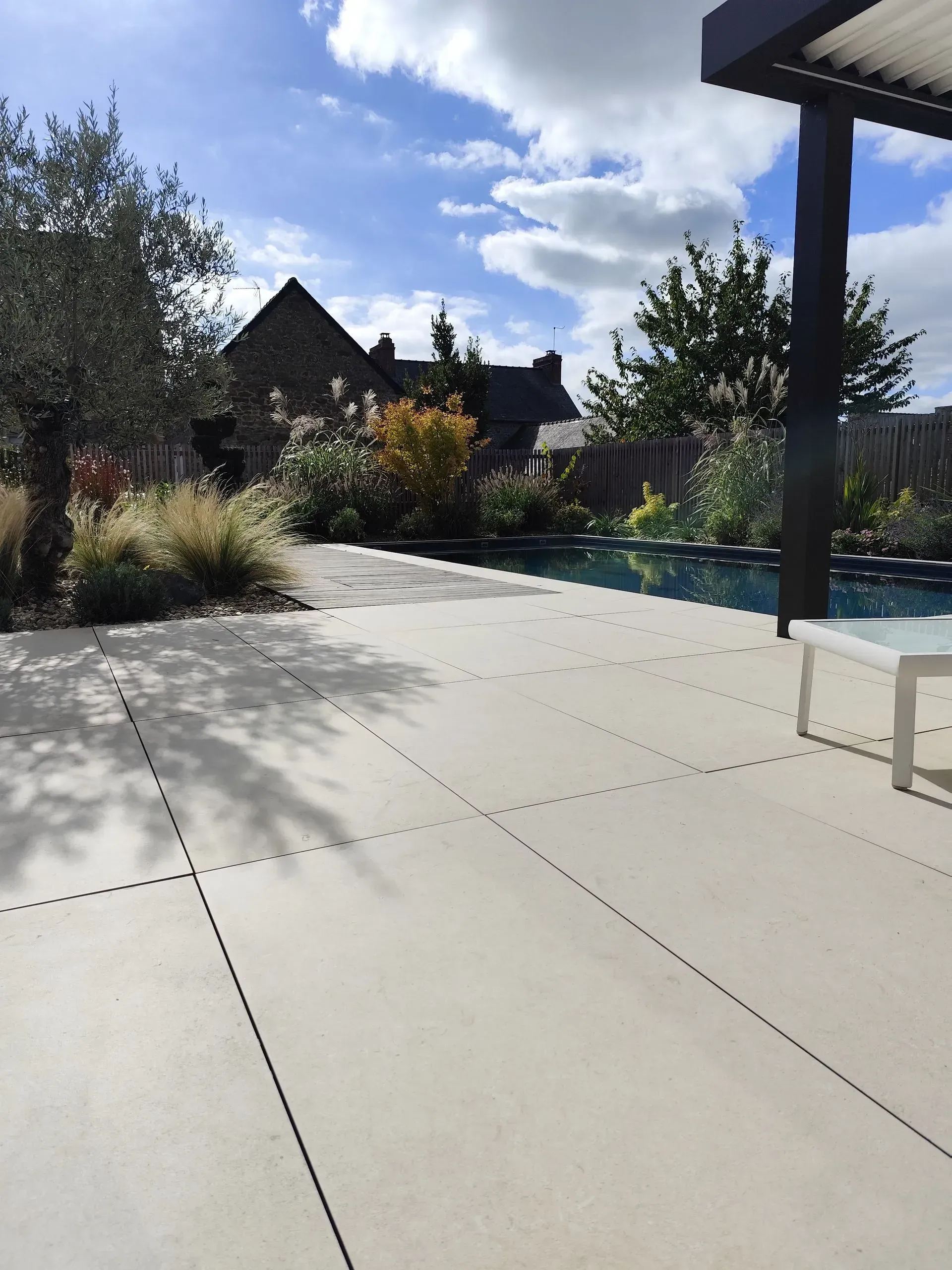 Patio ensoleillé dans la cour arrière avec dalles claires, piscine, plantes et maison sous un ciel bleu.