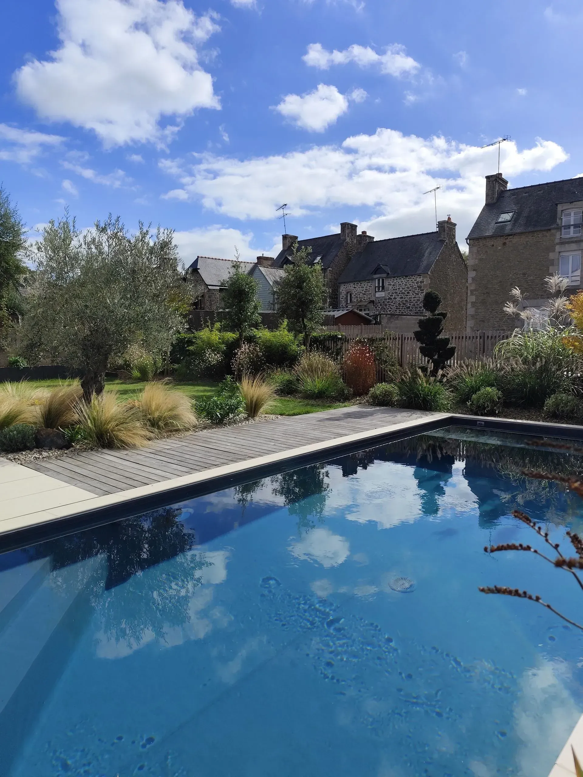 Piscine bleue à côté d'un jardin et de maisons en pierre sous un ciel lumineux, partiellement nuageux.