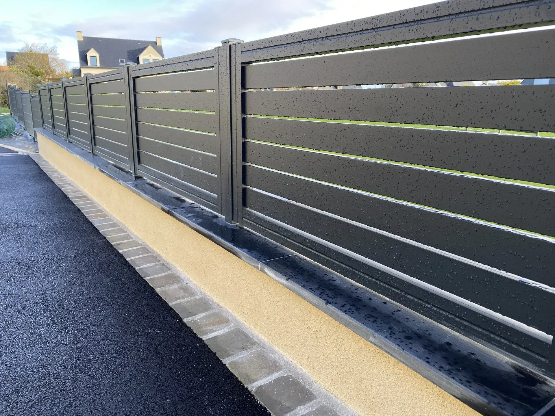Clôture grise à claire-voie le long d'une allée, bordée d'un trottoir jaune et d'une route asphaltée noire sous un ciel bleu.