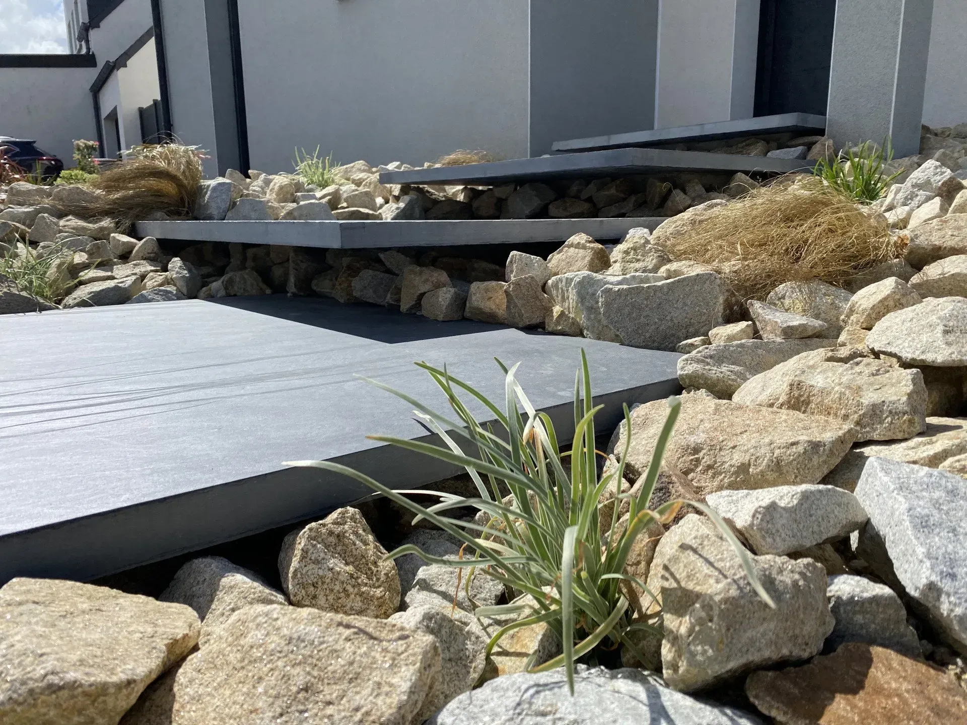 Jardin de rocaille jouxtant une terrasse couverte grise, agrémentée de plantes sèches et de pierres empilées.