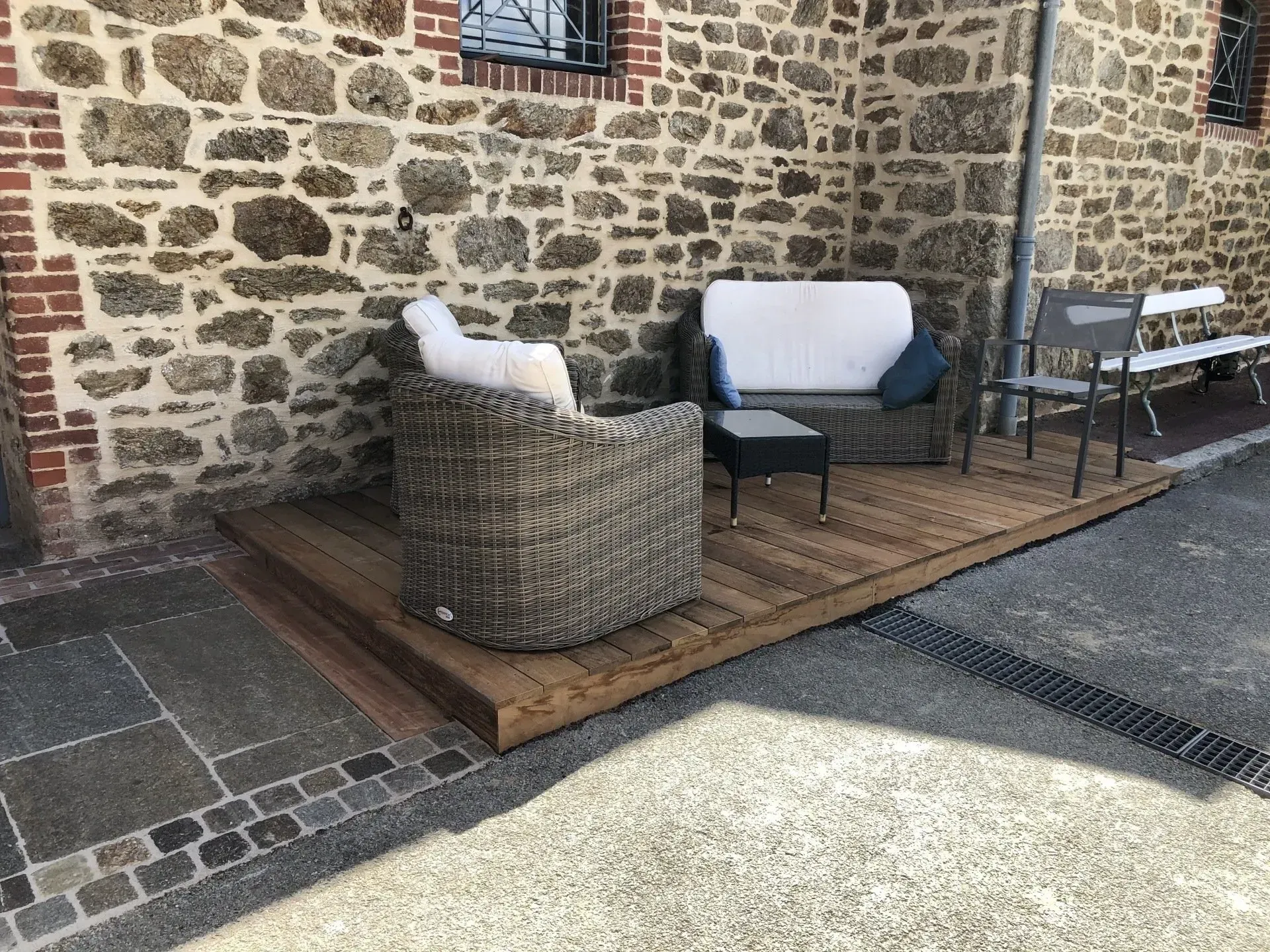 Terrasse en pierre avec fauteuil en osier, causeuse et petite table adossée à un mur en pierre.