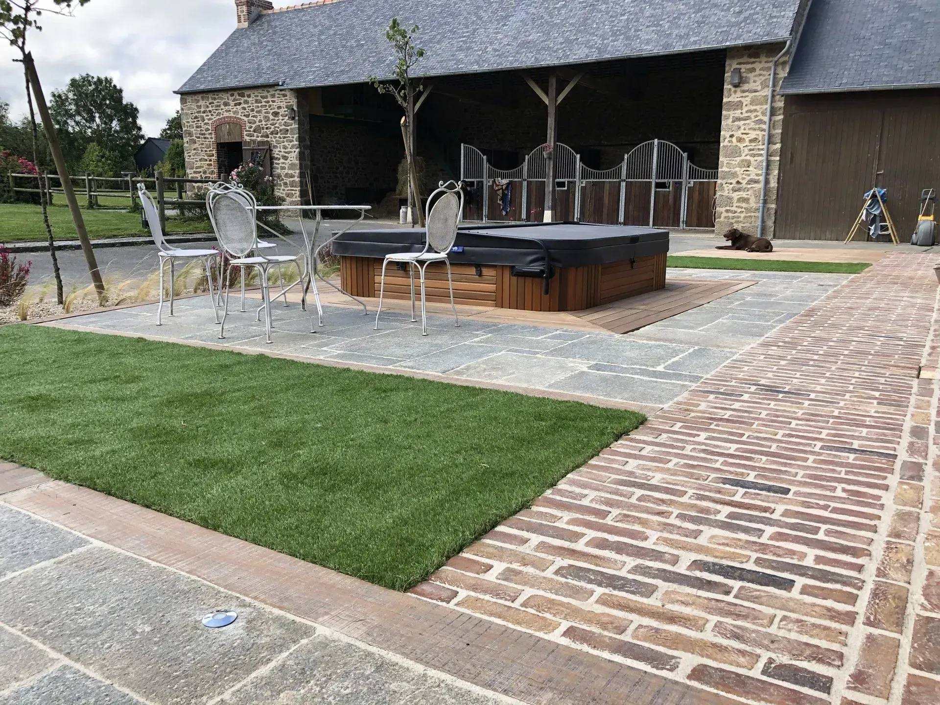 Terrasse en pierre avec jacuzzi, chaises en métal et allée en briques jouxtant un petit pavillon et une pelouse.