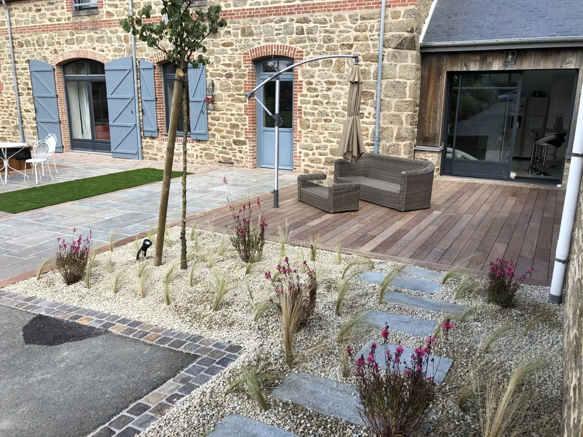 Petite cour avec bâtiment en pierre, terrasse en bois, bancs et quelques plantes éparses sur un sol en gravier.