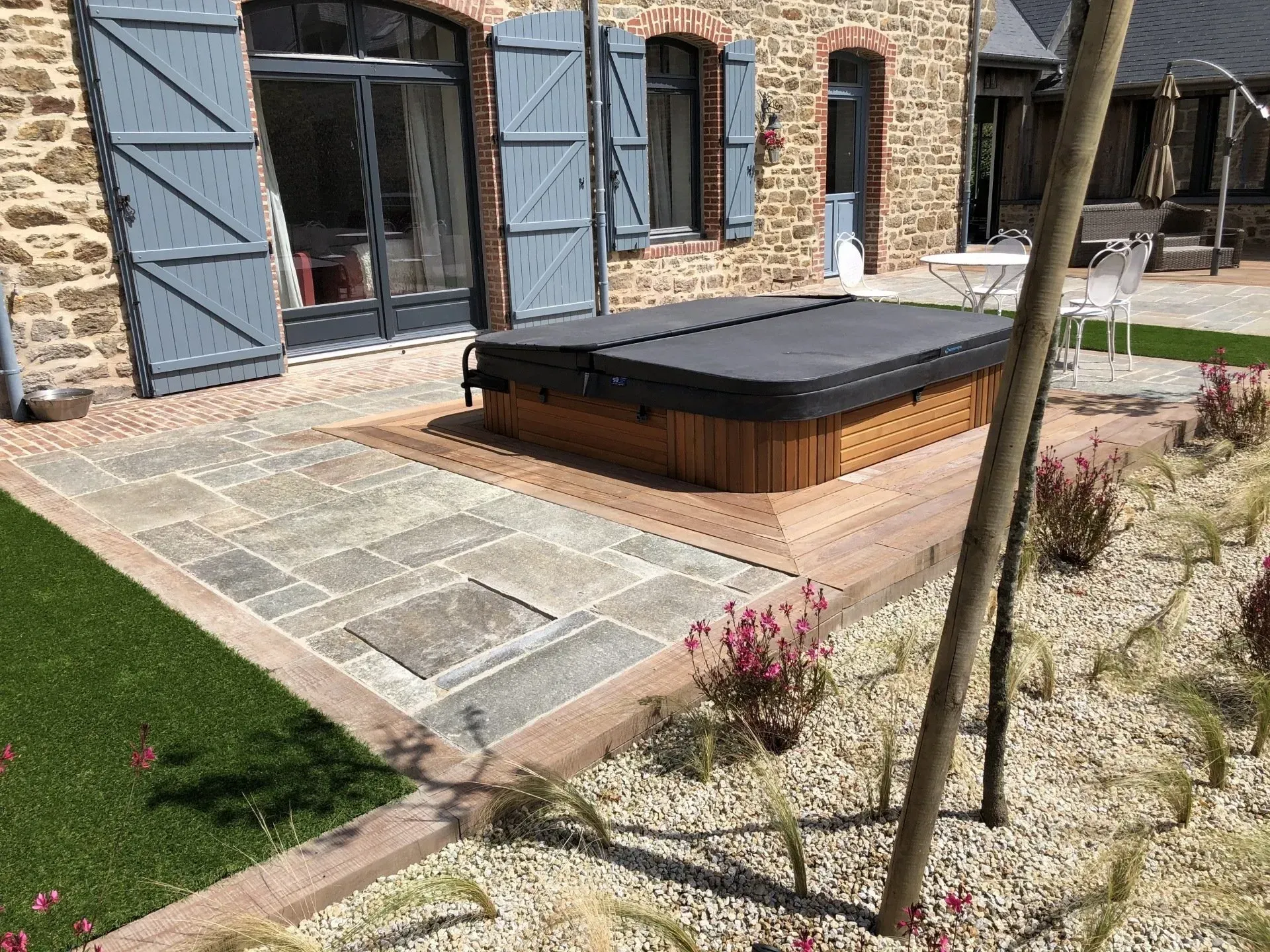 Terrasse en pierre avec un lit de jour extérieur à coussins noirs, jouxtant une maison en pierre et une pelouse.