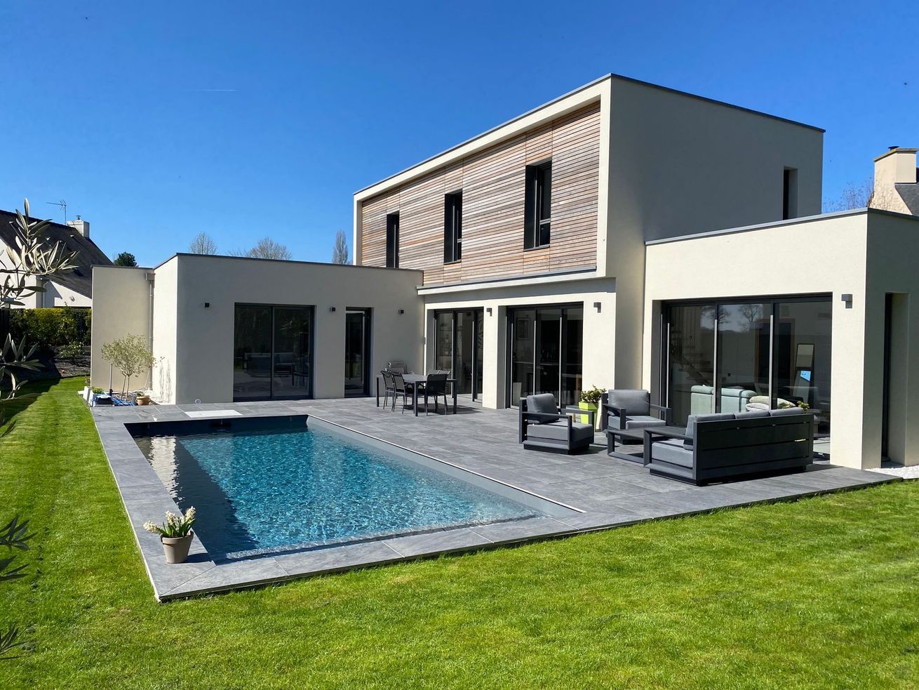 Maison moderne avec terrasse, piscine et pelouse verdoyante sous un ciel bleu azur.