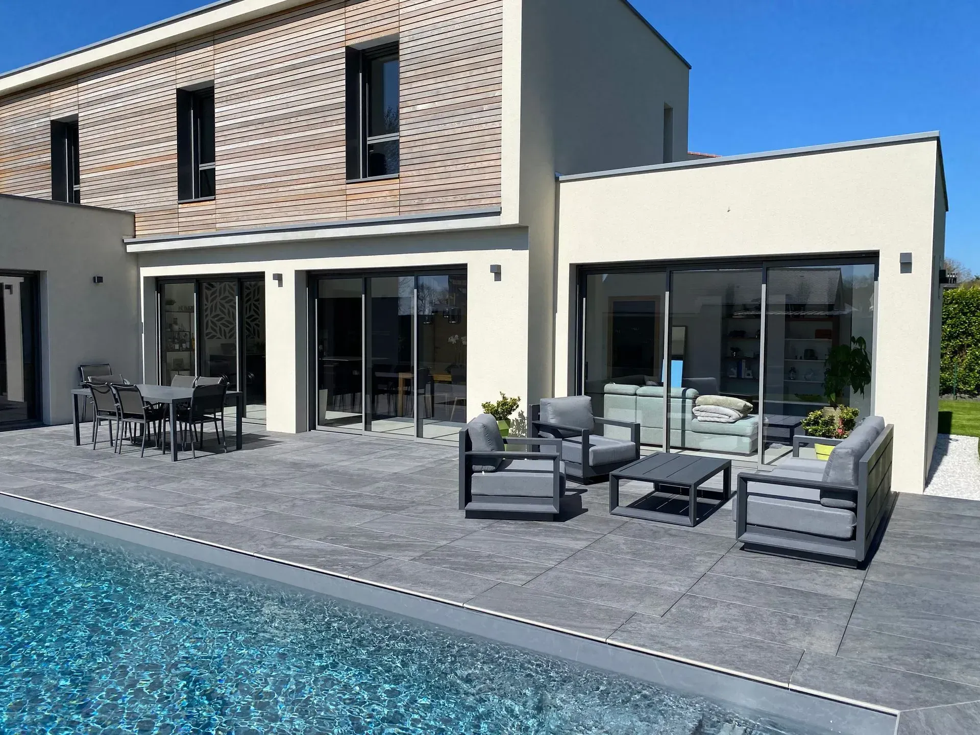 Patio moderne avec mobilier de jardin gris à côté d'une maison et d'une piscine sous un ciel bleu azur.