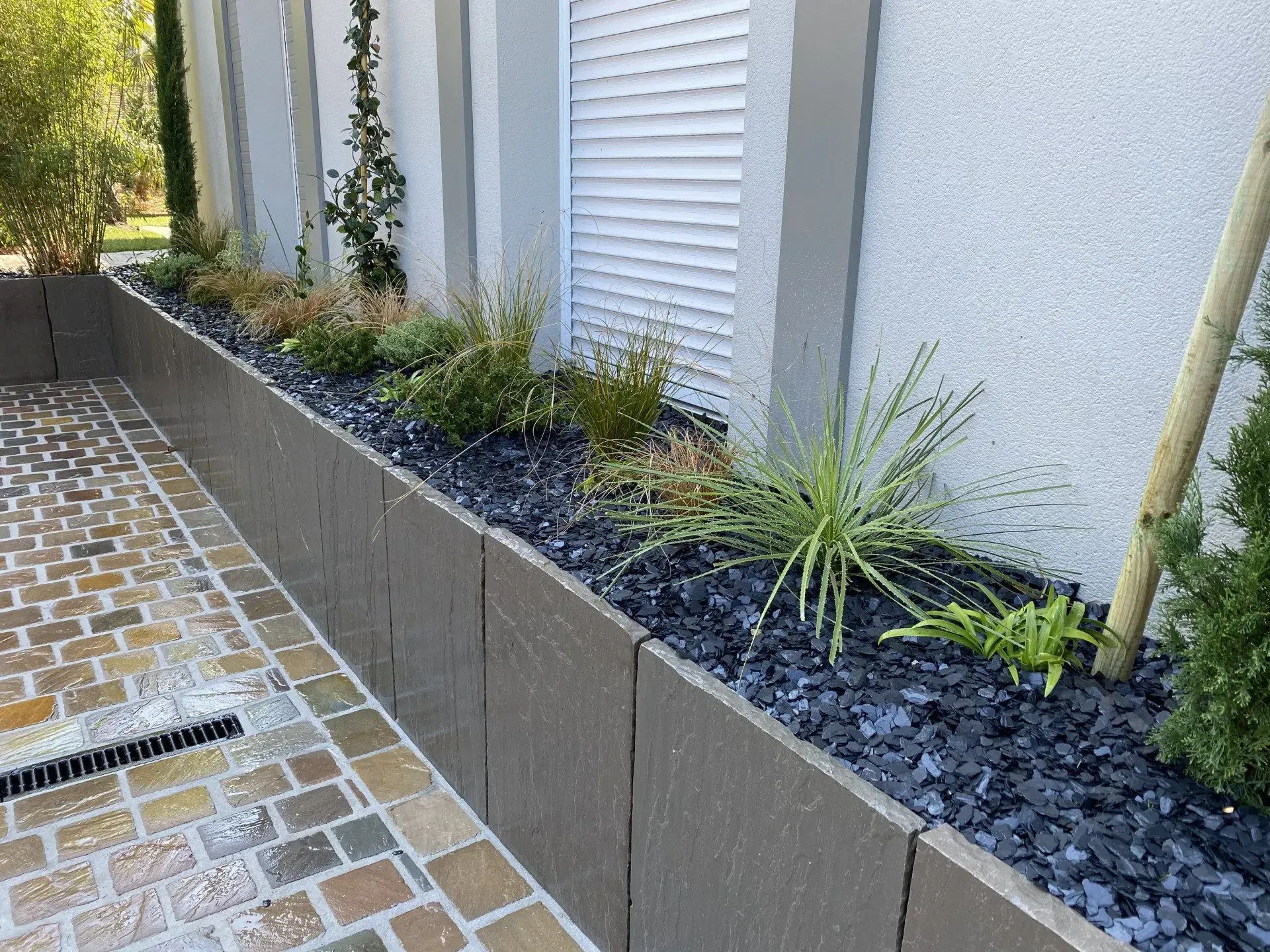 Allée moderne longeant un bâtiment blanc, agrémentée d'une jardinière surélevée recouverte de paillis noir et de petites plantes vertes.