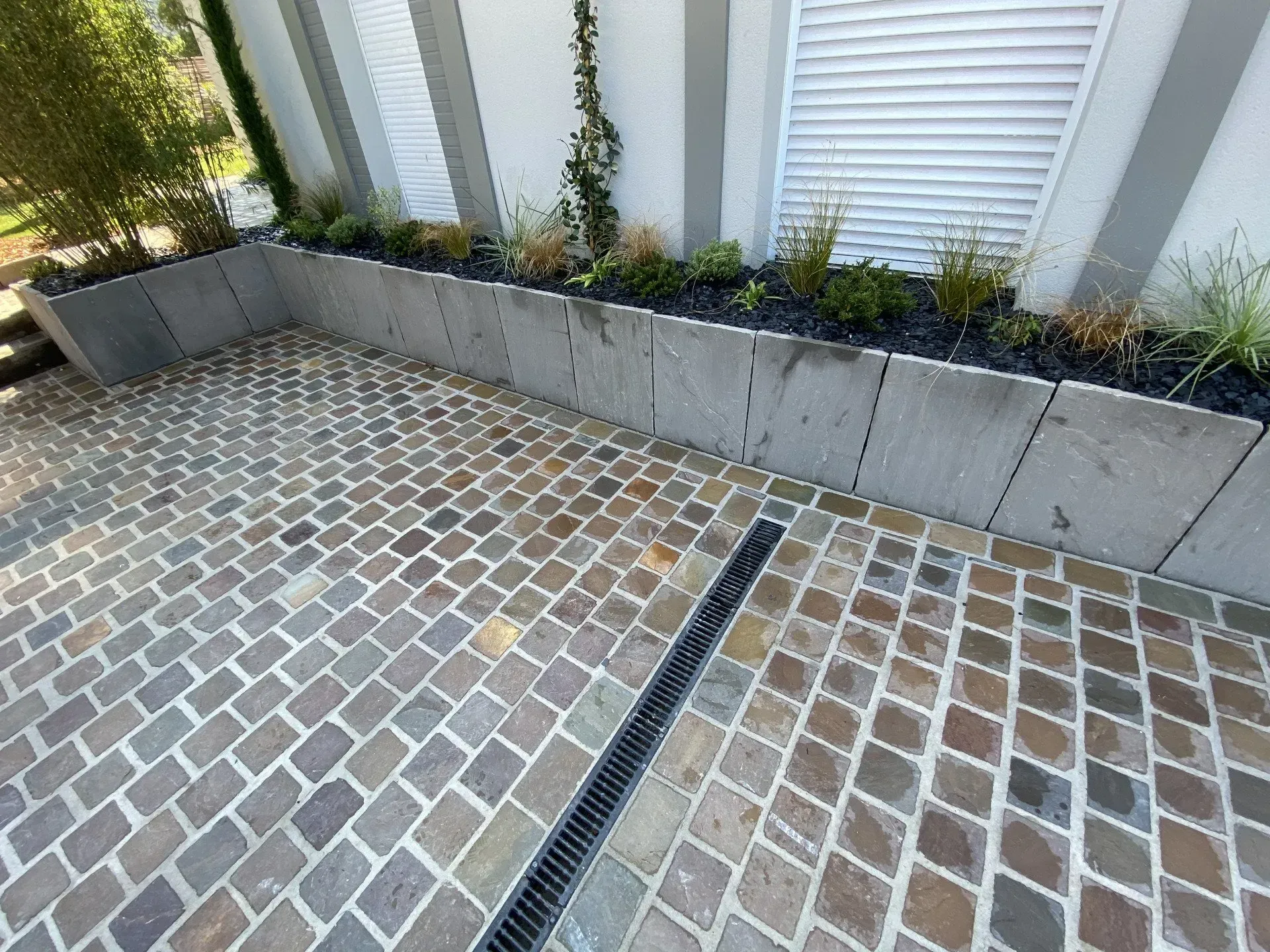 Terrasse pavée de briques avec jardinières en béton et volets blancs le long d'un mur.