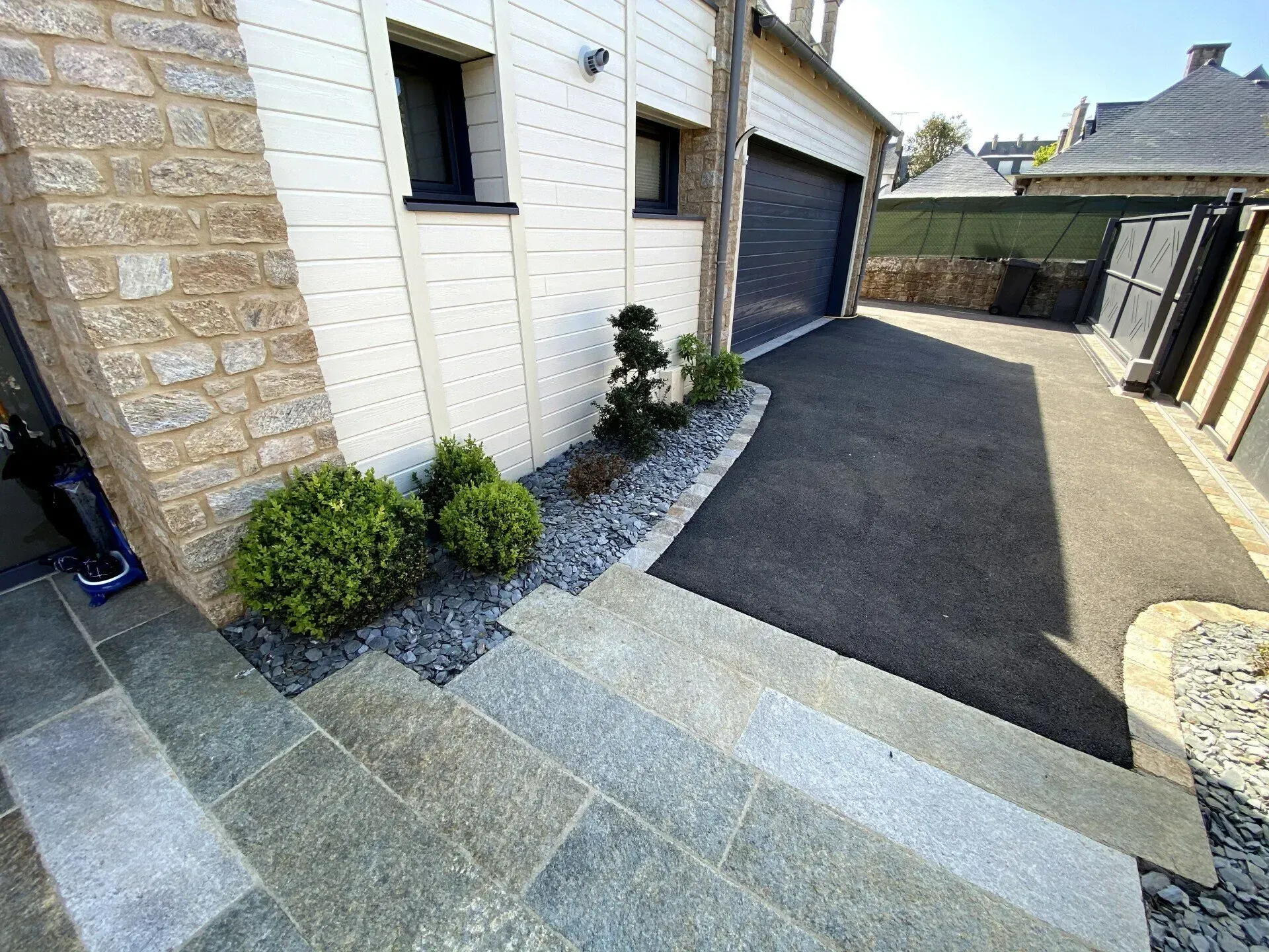 Allée d'accès à une maison avec pavage à motifs, asphalte noir, porte de garage et petits arbustes le long du mur.