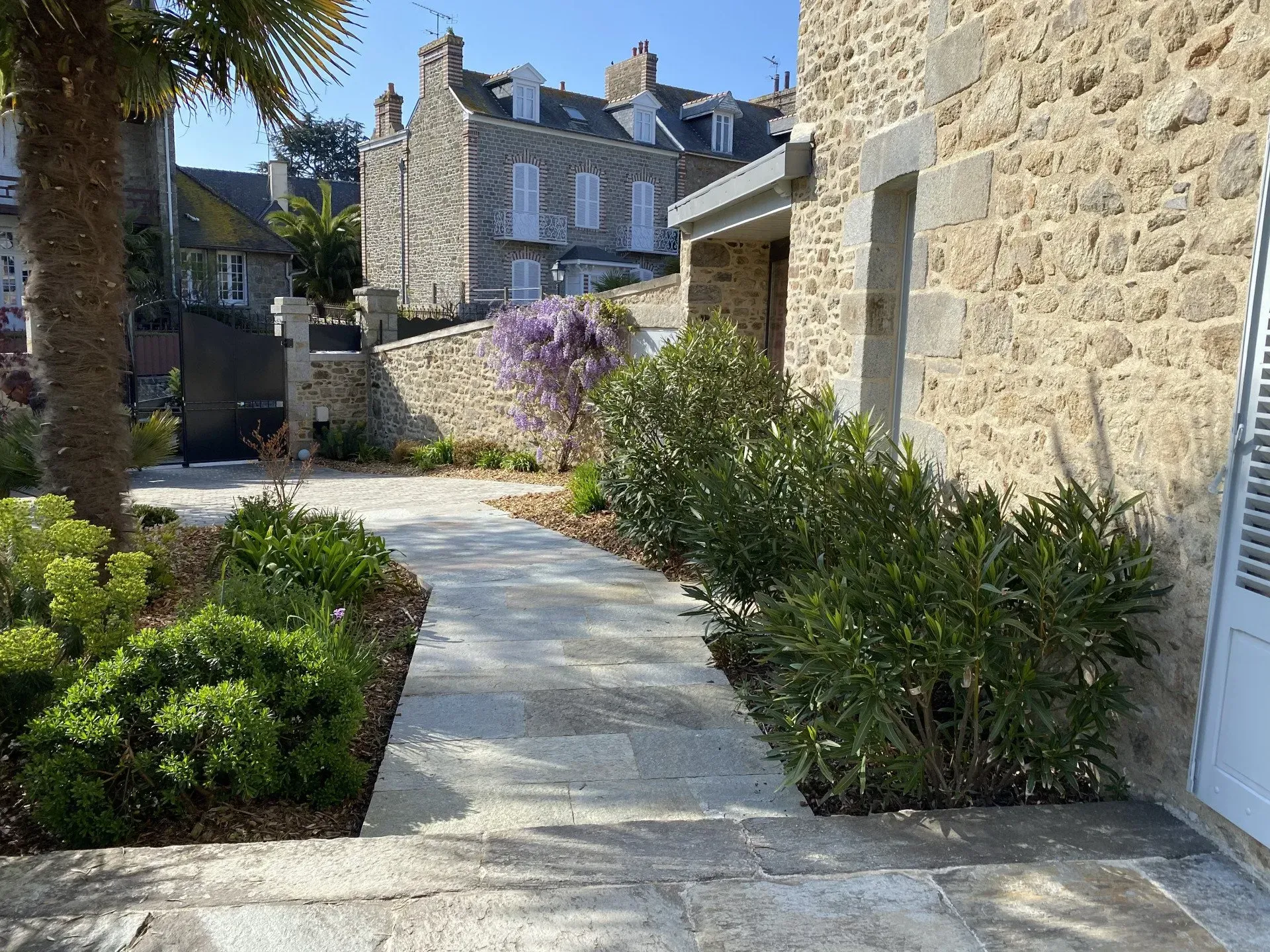 Allée de pierre traversant une cour paysagée, bordée de maisons en pierre, sous un ciel d'un bleu limpide.