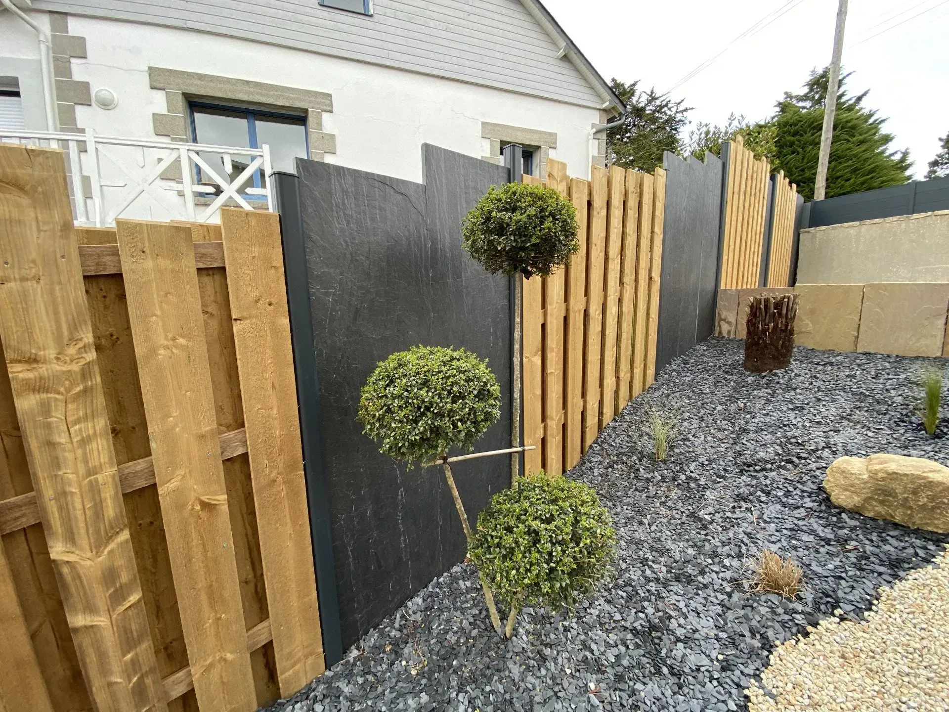 Jardin moderne avec allée de gravier, clôture en bois, portail noir et arbres topiaires taillés.