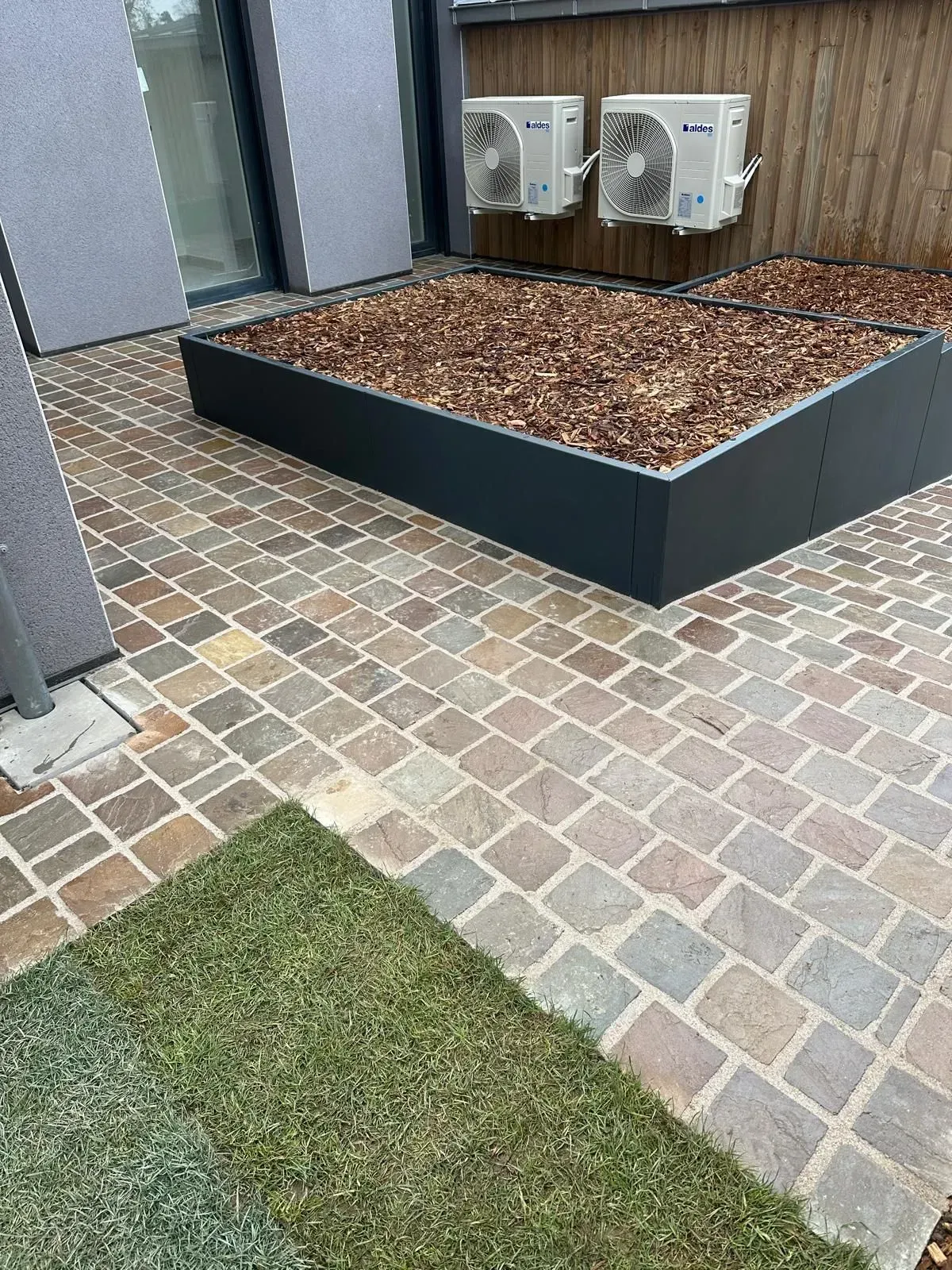 Terrasse extérieure avec pavés de briques claires, gazon artificiel, jardinière surélevée et unités de climatisation murales.