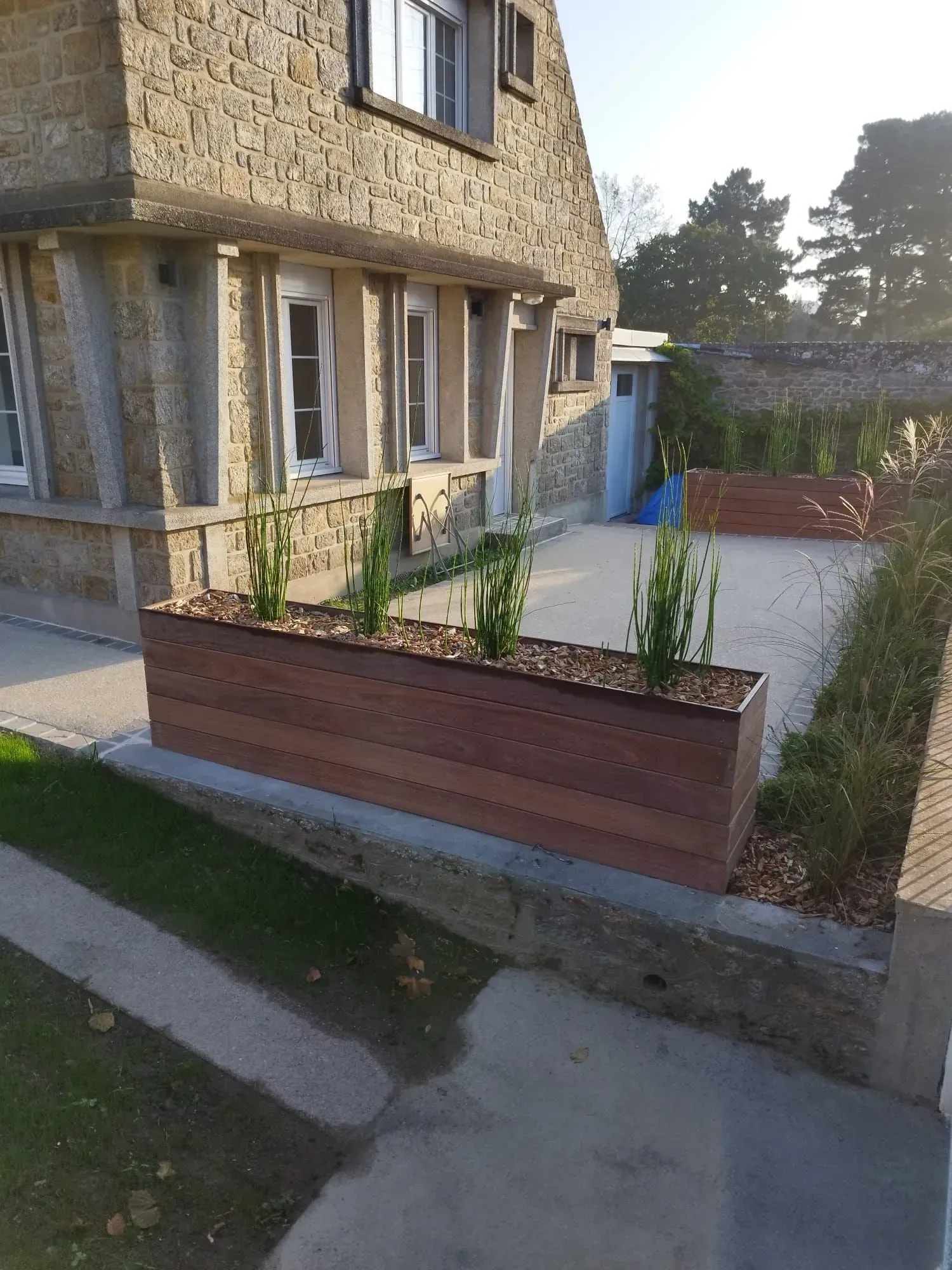 Entrée d'un bâtiment en pierre avec des parterres surélevés et une allée pavée menant à un portail bleu.