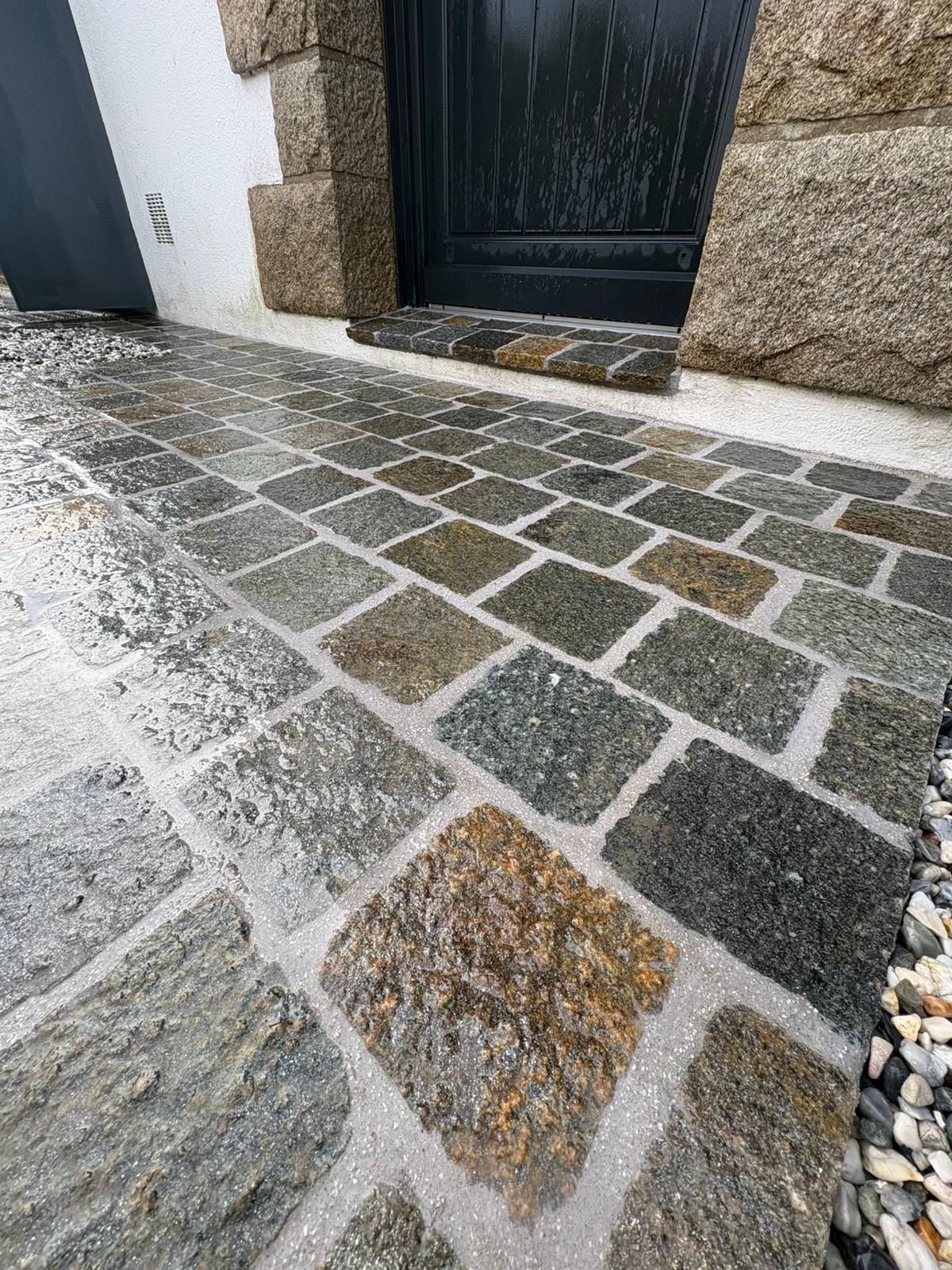 Allée en pierre mouillée menant à une porte noire située à côté d'un mur texturé et d'une bordure de galets.