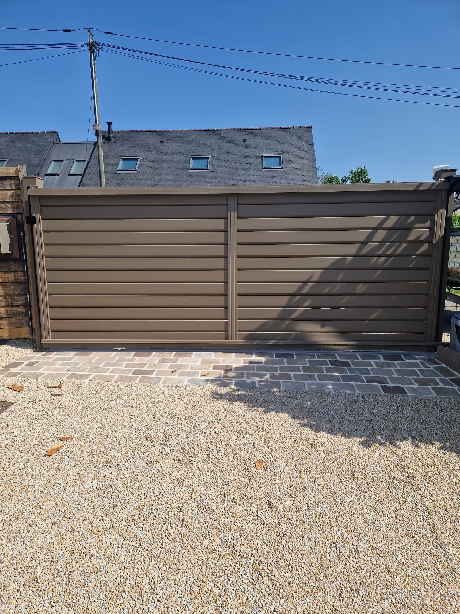 Portes de garage en métal brun sur une allée de gravier, avec une maison et un ciel bleu en arrière-plan.