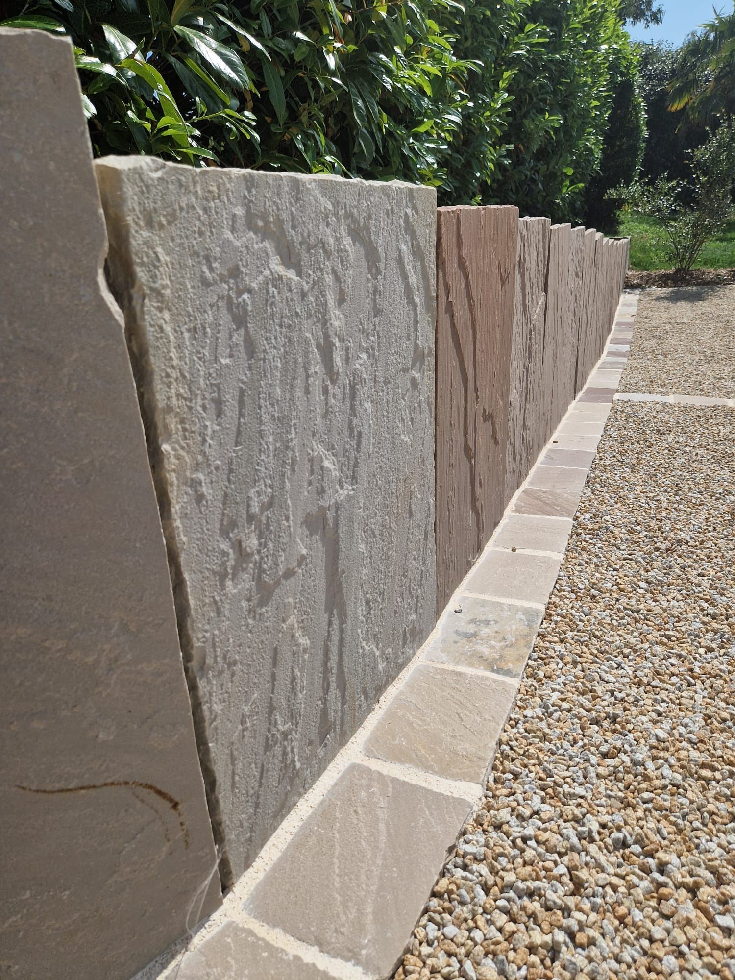 Mur de soutènement en pierre bordant un chemin de gravier, avec des arbres verts en arrière-plan.
