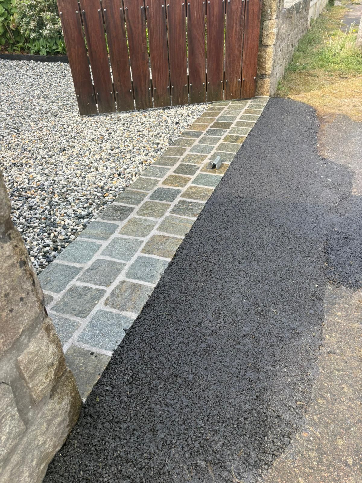 Chemin de gravier longeant un mur de pierre, bordé d'un étroit pavage et d'une allée ou d'un trottoir sombre.