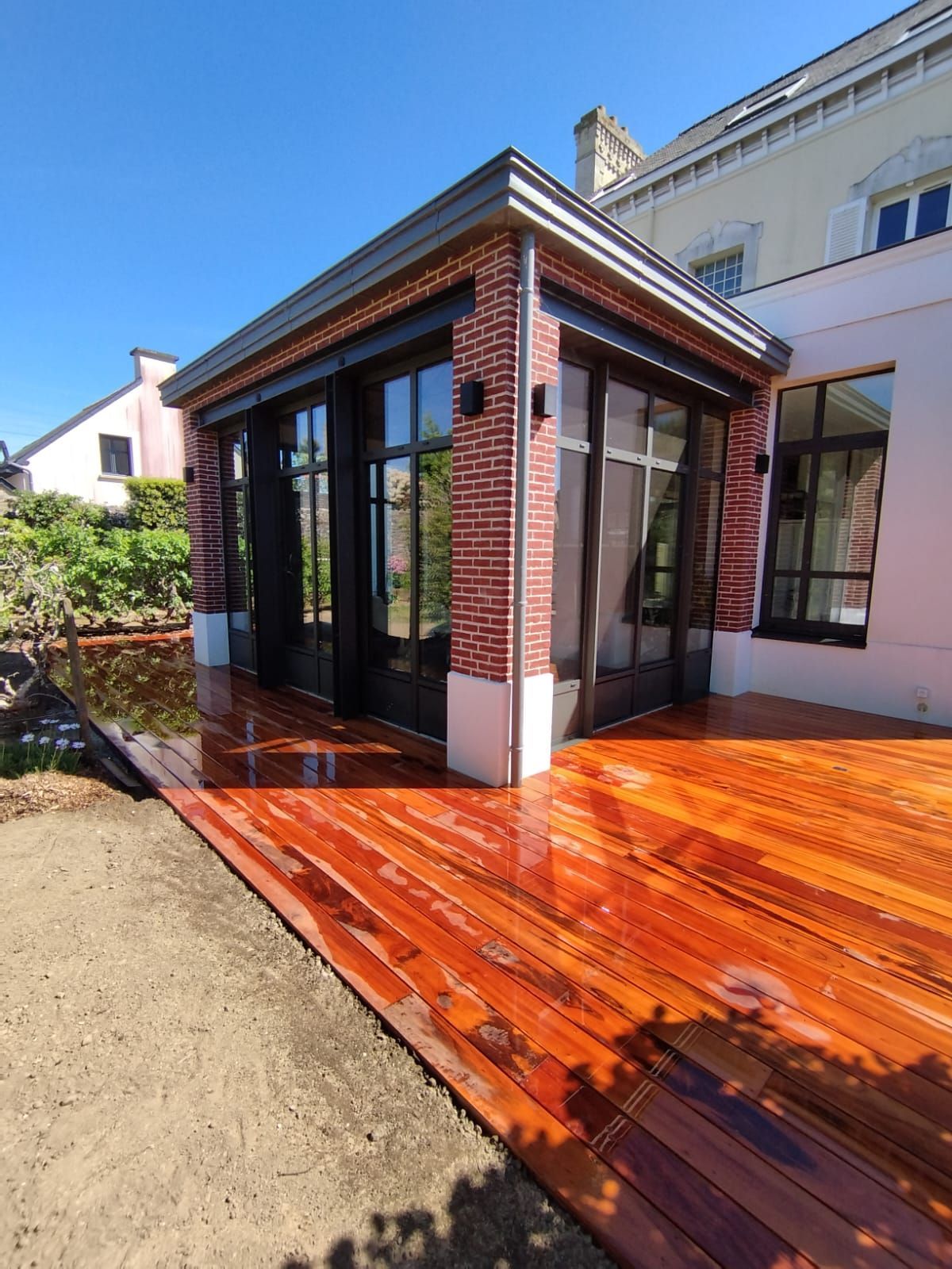 Véranda moderne avec parois vitrées et terrasse en bois rouge brillant donnant sur une cour gravillonnée.