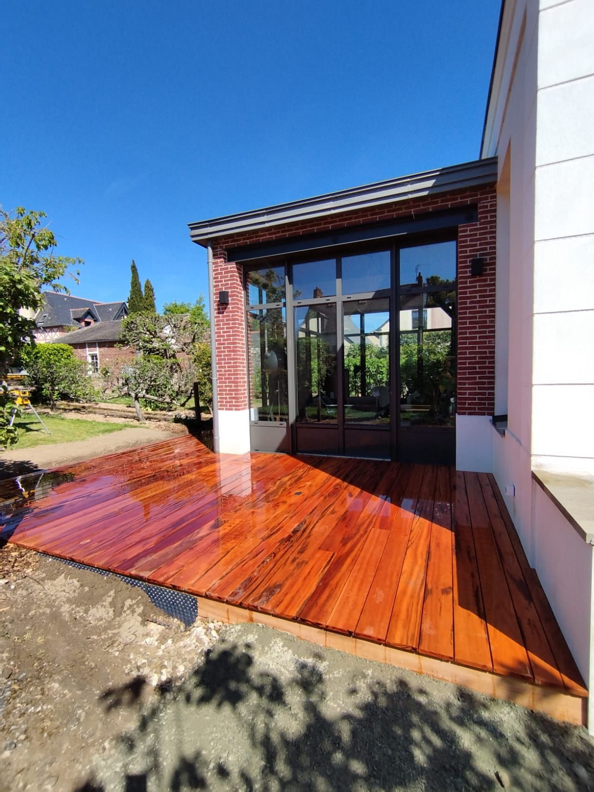 Terrasse en bois verni jouxtant une maison aux portes coulissantes en verre, sous un ciel bleu.