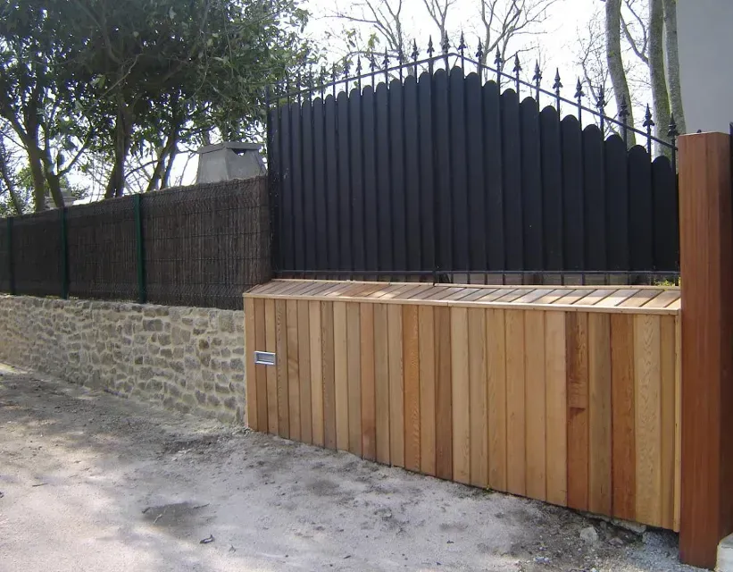 Clôture et portail en bois jouxtant un mur de pierre le long d'une allée bordée d'arbres.