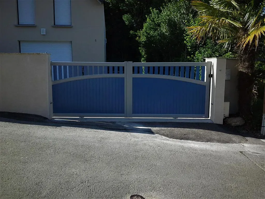 Portail d'allée en métal bleu à côté d'une maison beige, avec du soleil et des arbres en arrière-plan.