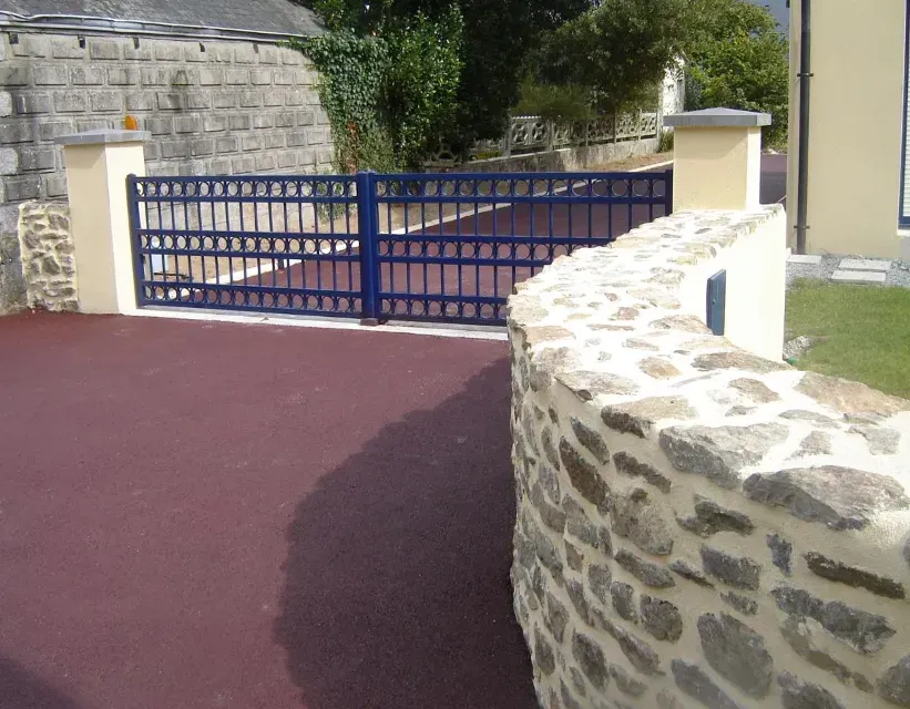 Portail en métal bleu et mur de pierre à l'entrée d'une cour pavée de rouge.