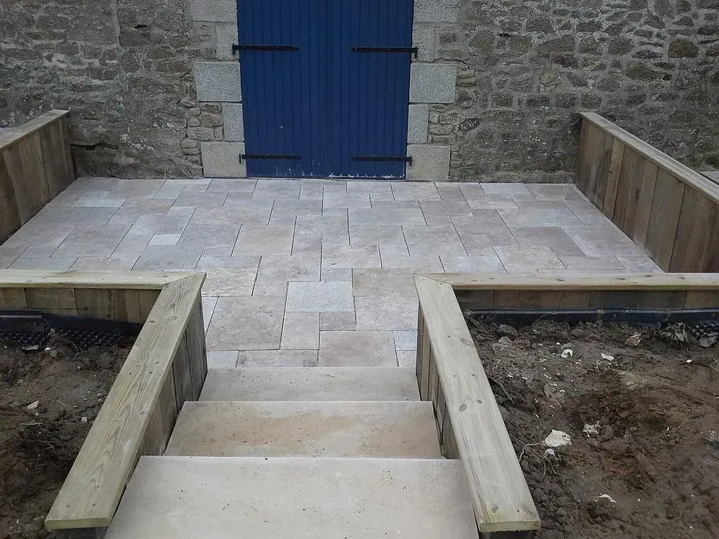 Des marches en pierre mènent à une porte bleue, avec une balustrade en bois basse et un palier pavé.