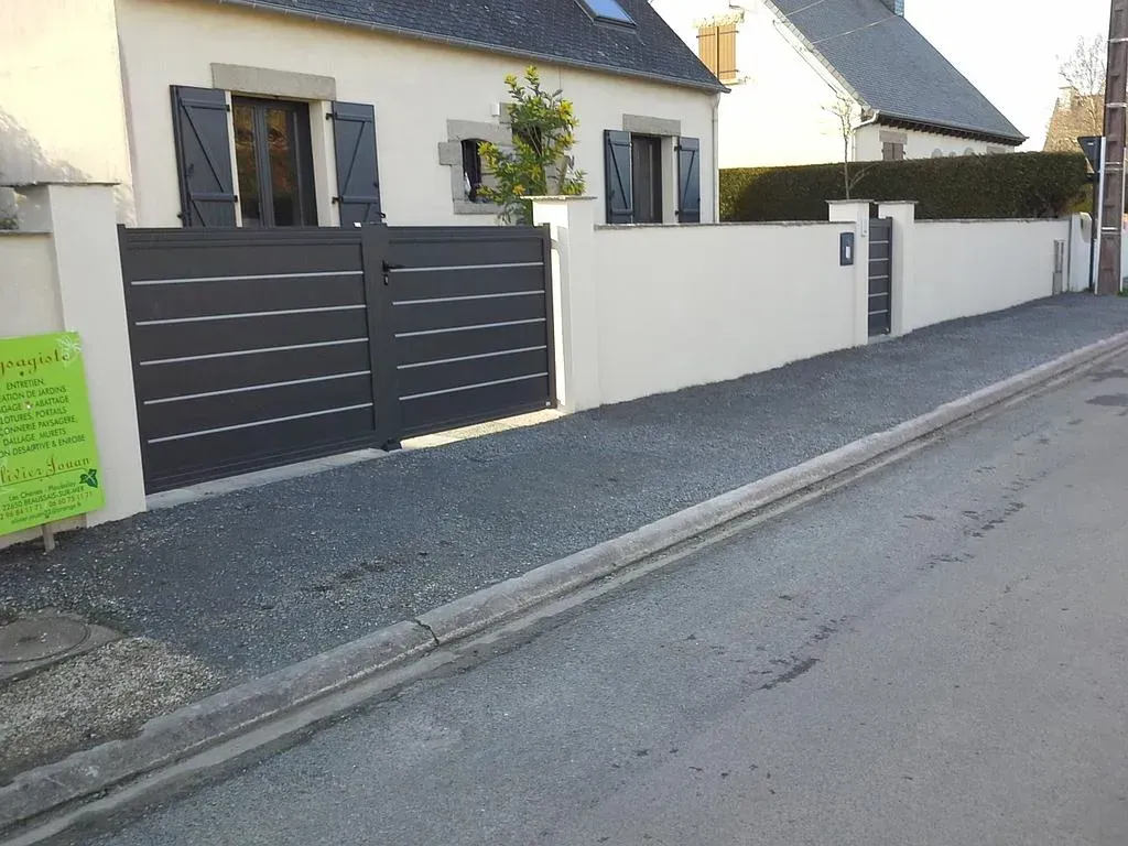 Maison d'habitation avec portail gris et mur blanc, située le long d'une rue pavée tranquille.