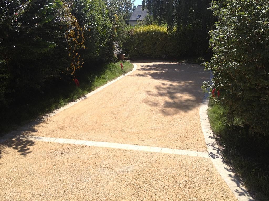 Allée de gravier ensoleillée bordée de haies, avec une ligne blanche en travers.