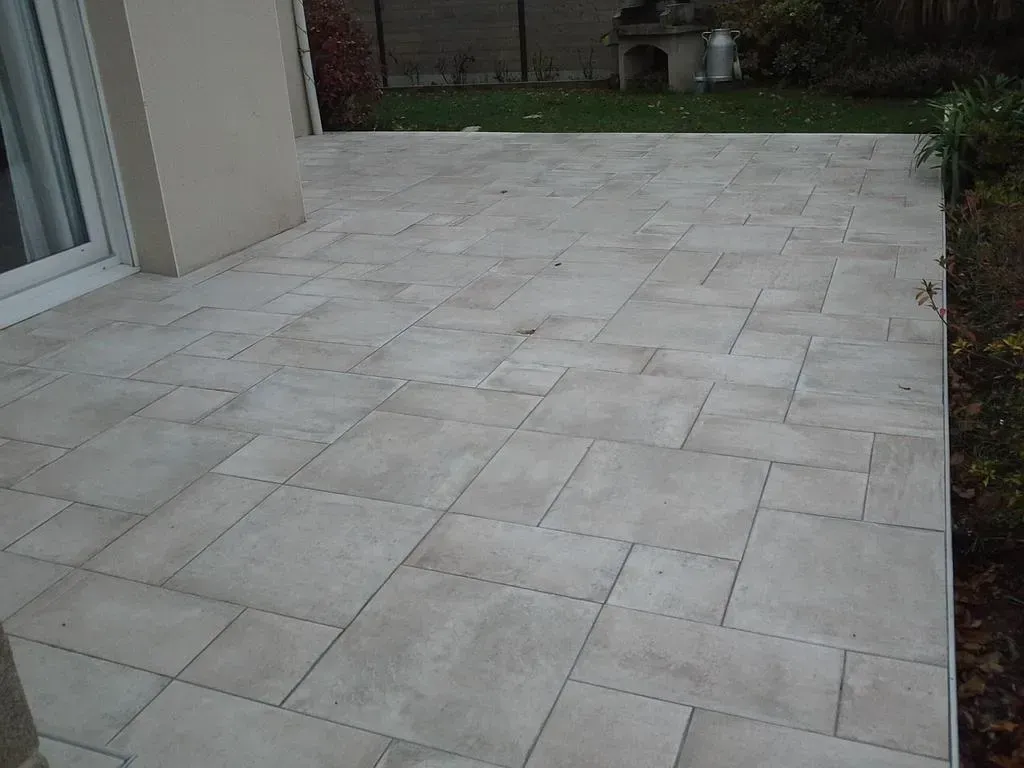 Terrasse en pierre avec pavés gris clair, jouxtant une maison et un chemin de jardin, vue depuis une porte.